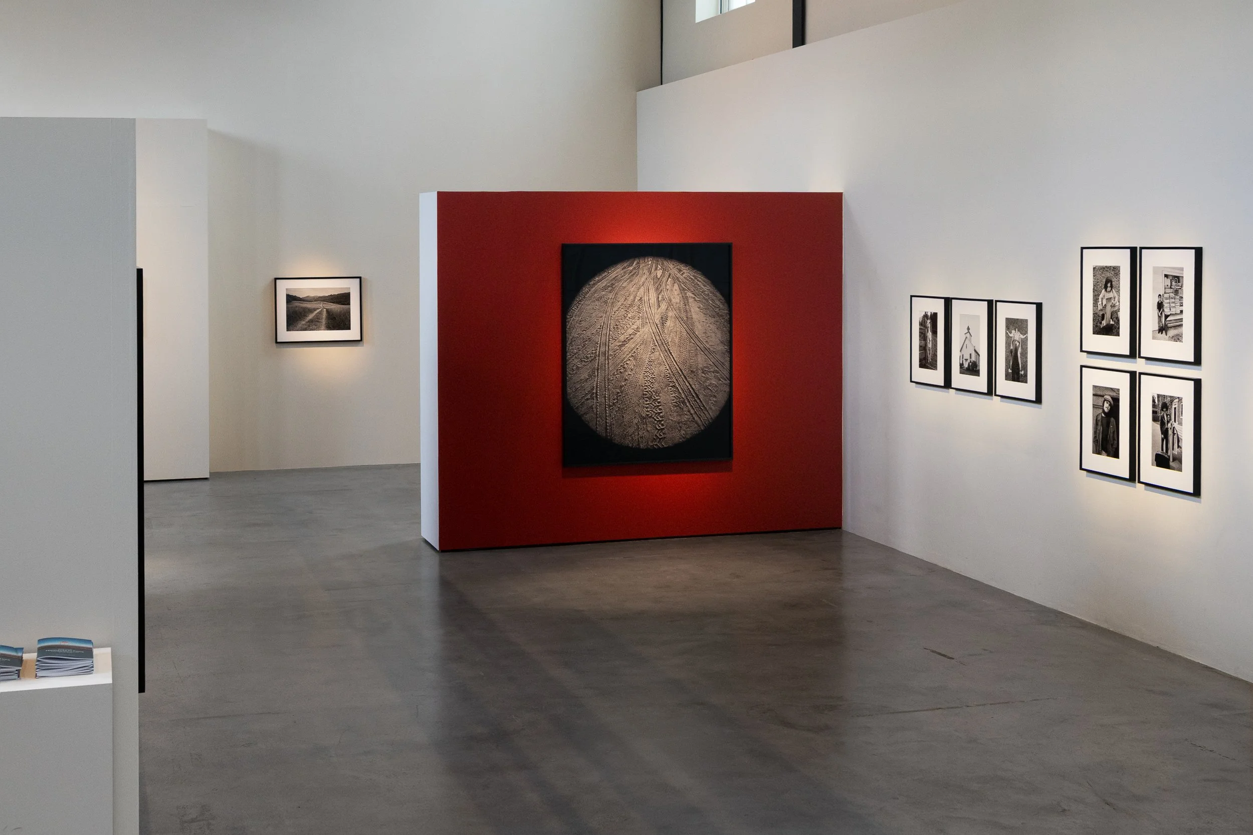 An art gallery with black and white photographic images on white walls, featuring a red-paneled wall with a large circular artwork in the center, gray flooring, and a high ceiling with large windows.
