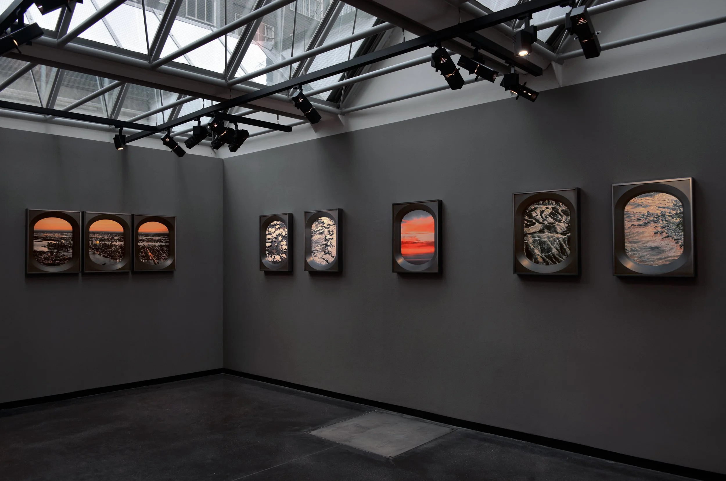 An art gallery with framed landscape photographs on a gray wall, a large skylight, and multiple ceiling-mounted black spotlights.