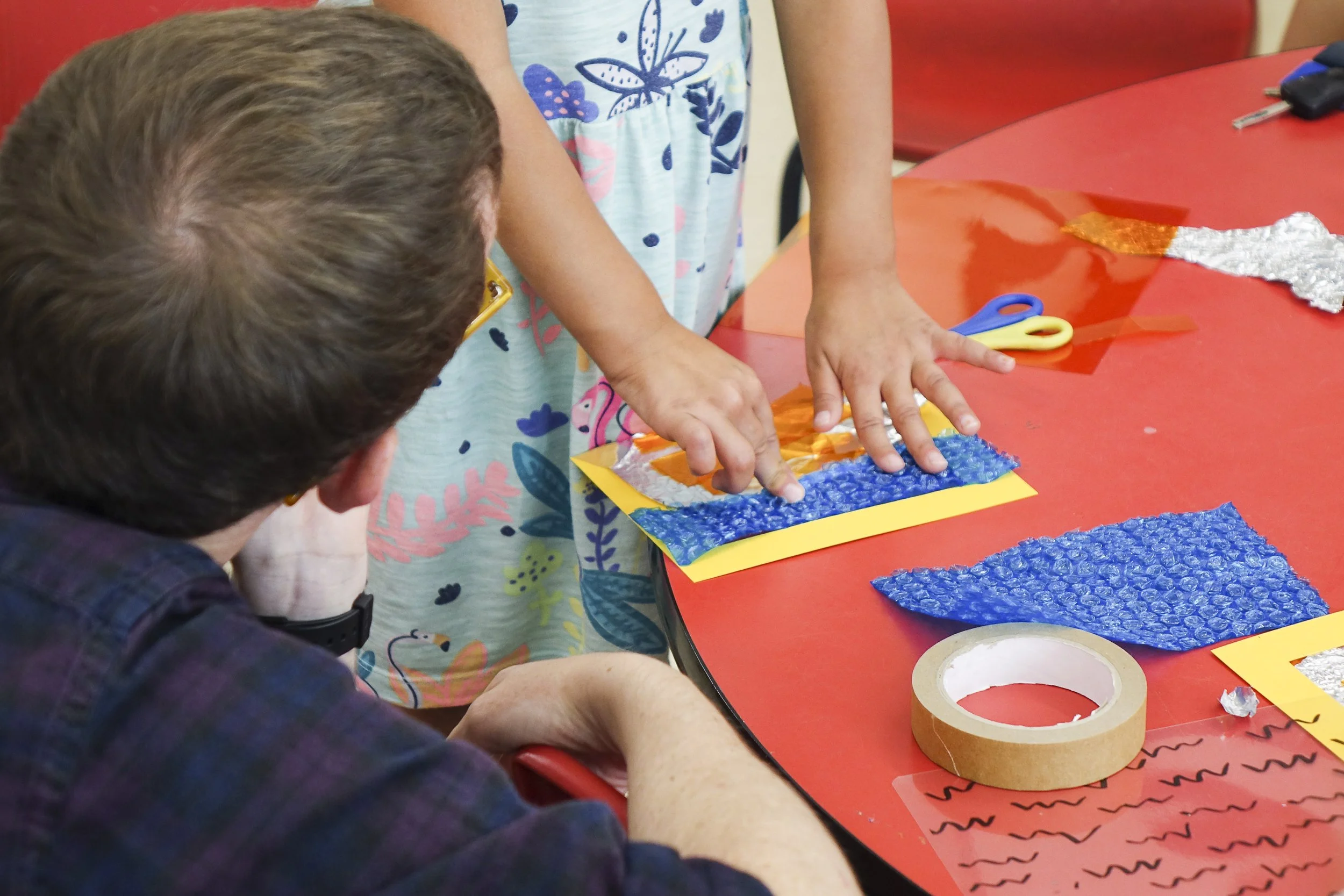 Two children are working on a craft project at a red table with various craft materials like foam sheets, tape, scissors, and decorative foil.