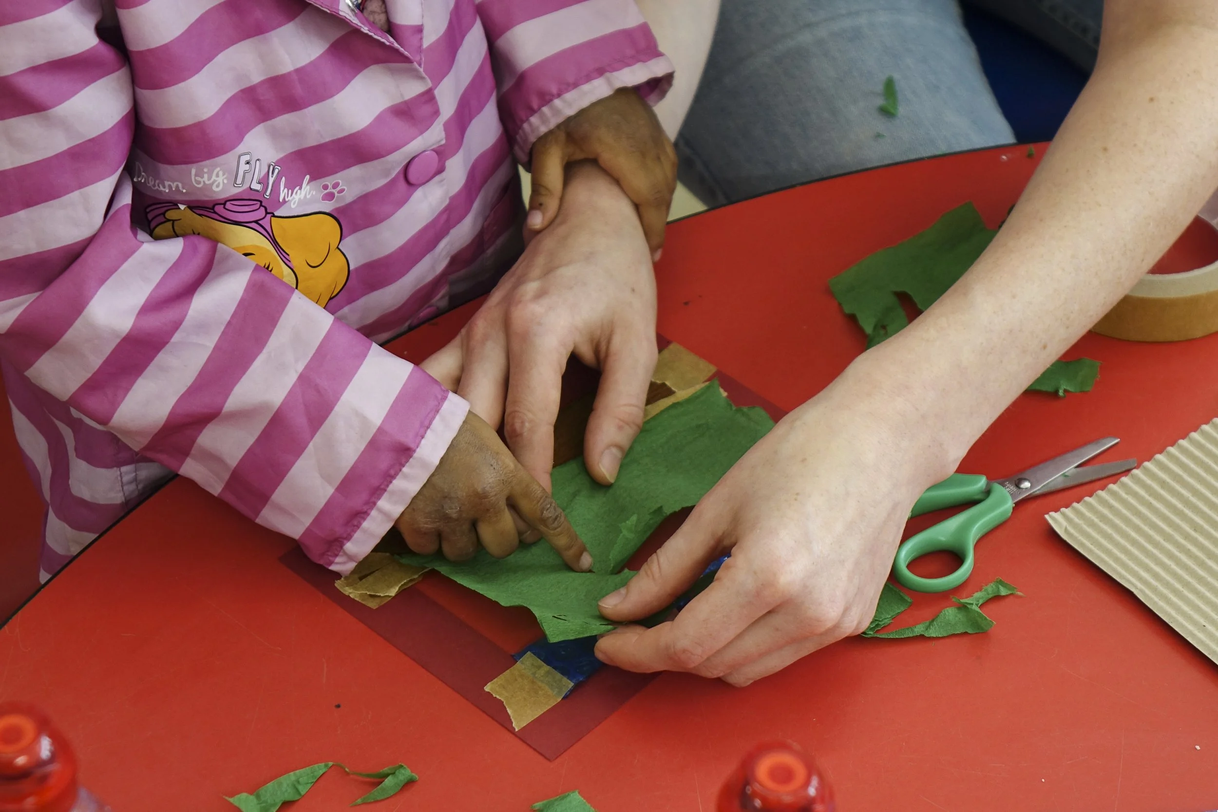 Multiple hands working together to cut and arrange green tissue paper on a red surface for crafting, with scissors and tape nearby.