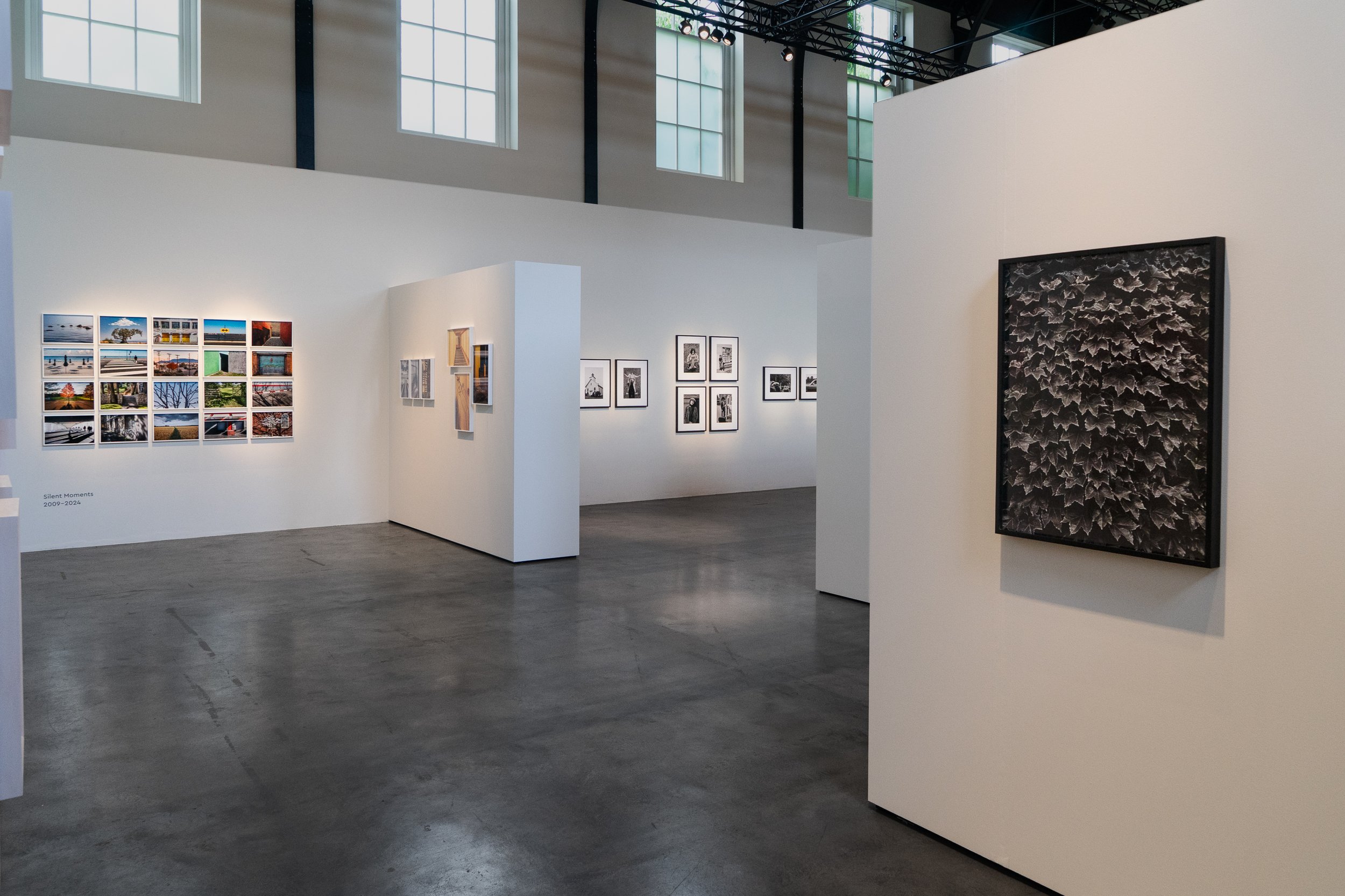 Interior view of an art gallery with white walls displaying photos and artwork, large windows, and a high ceiling with lighting fixtures.