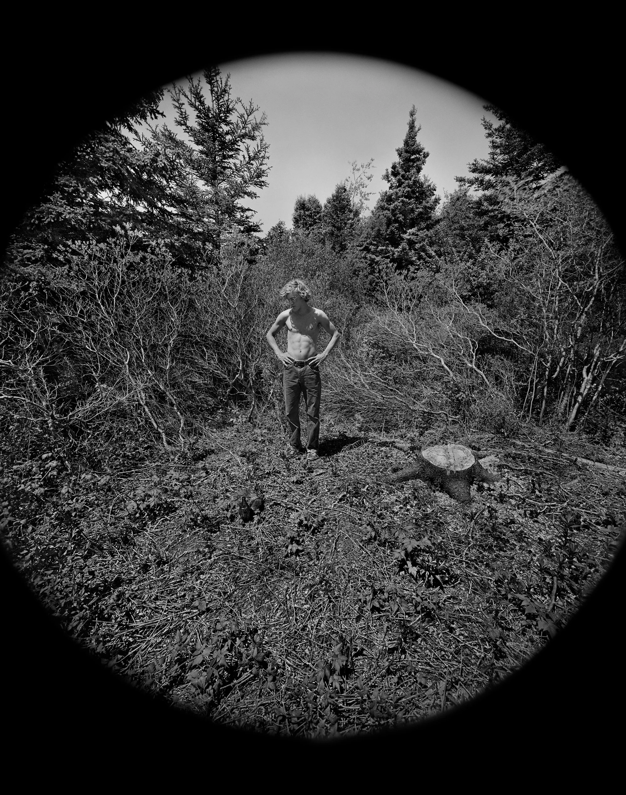 A black and white photo of a shirtless young man with curly hair standing in a wooded area, viewed through a circular opening.