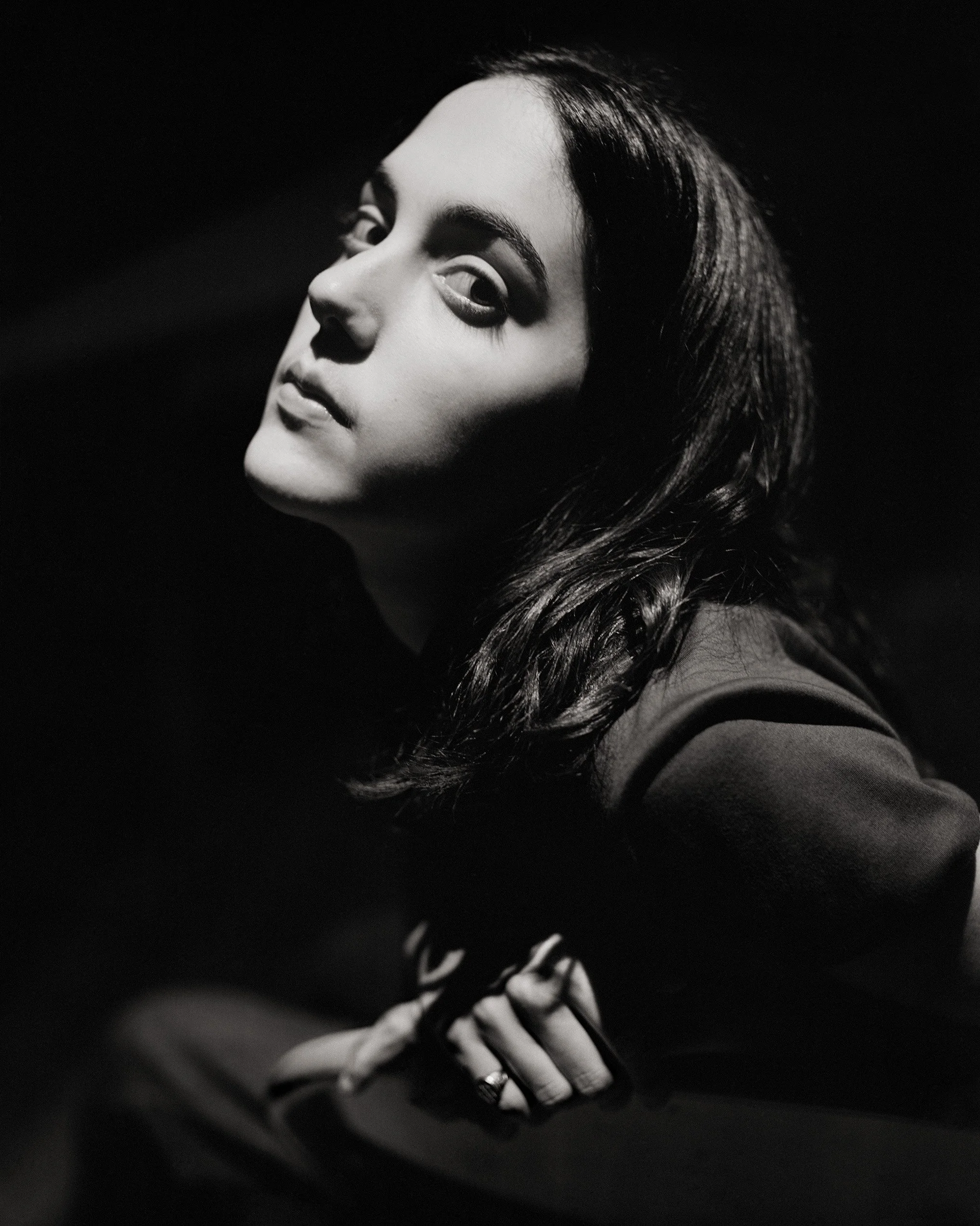 Black and white photograph of a young woman with dark hair and strong eyebrows, looking over her shoulder, with part of her face illuminated against a dark background, and her hands resting on her lap.
