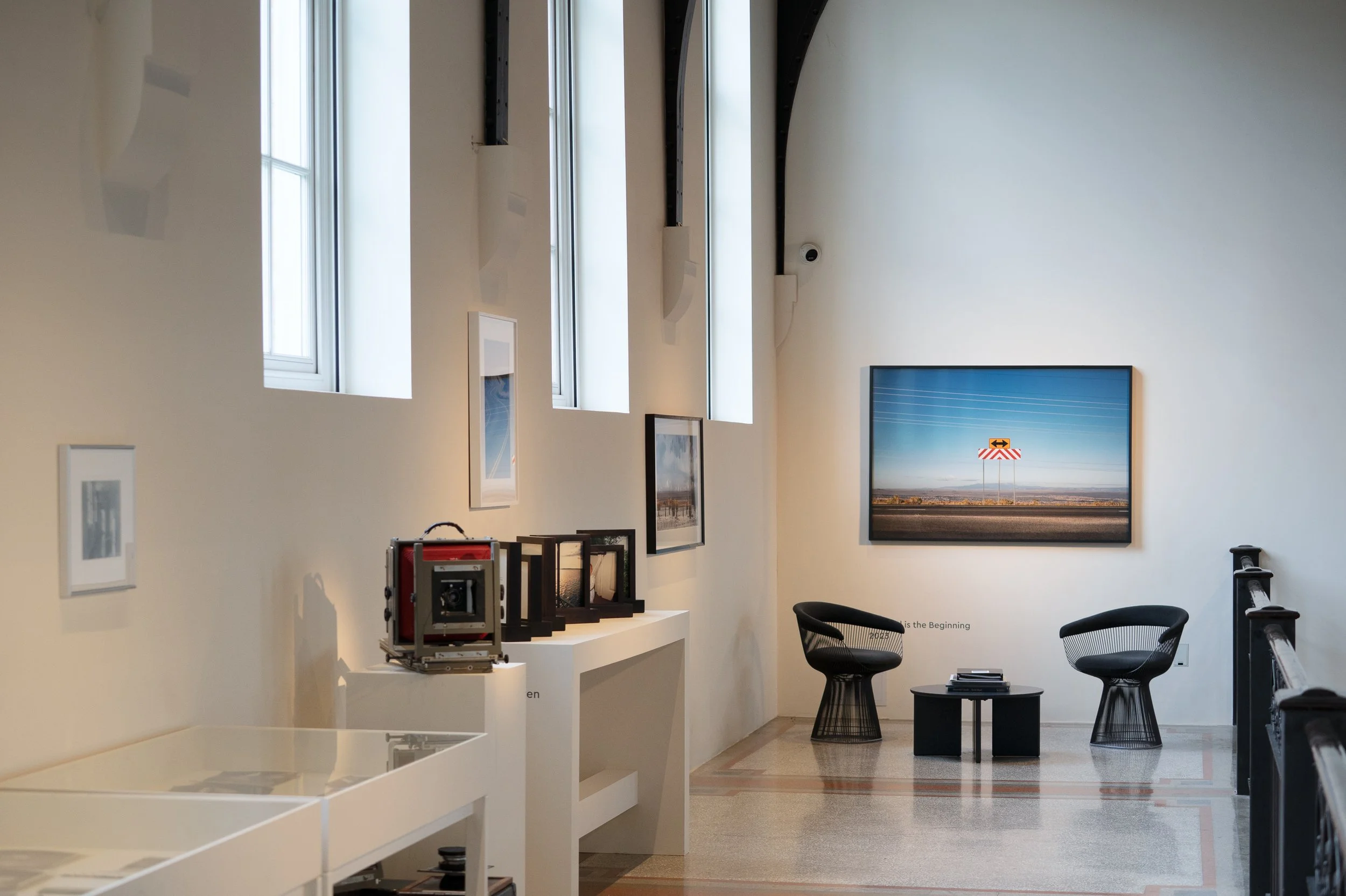 An art gallery with framed photographs on the wall, a display table with a vintage camera, and modern chairs.