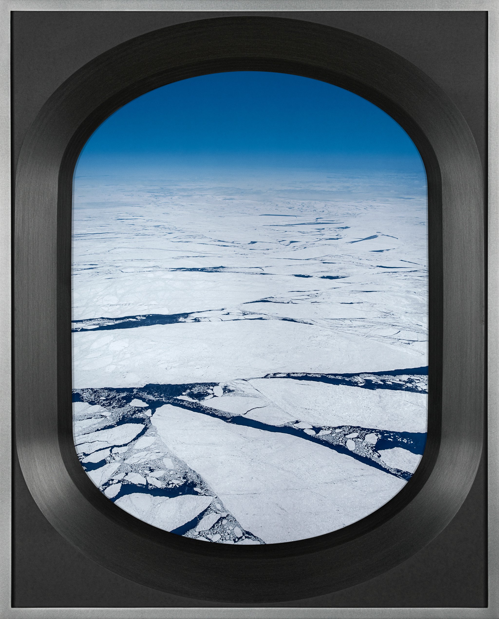 View through an airplane window of an icy landscape with fractured ice and a clear blue sky.