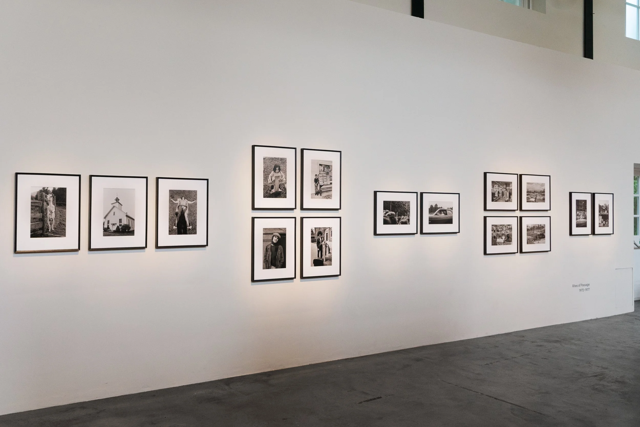 Gallery wall displaying black-and-white photographs in frames, labeled "Rites of Passage 1972-1977," in an art gallery with white walls and gray flooring.