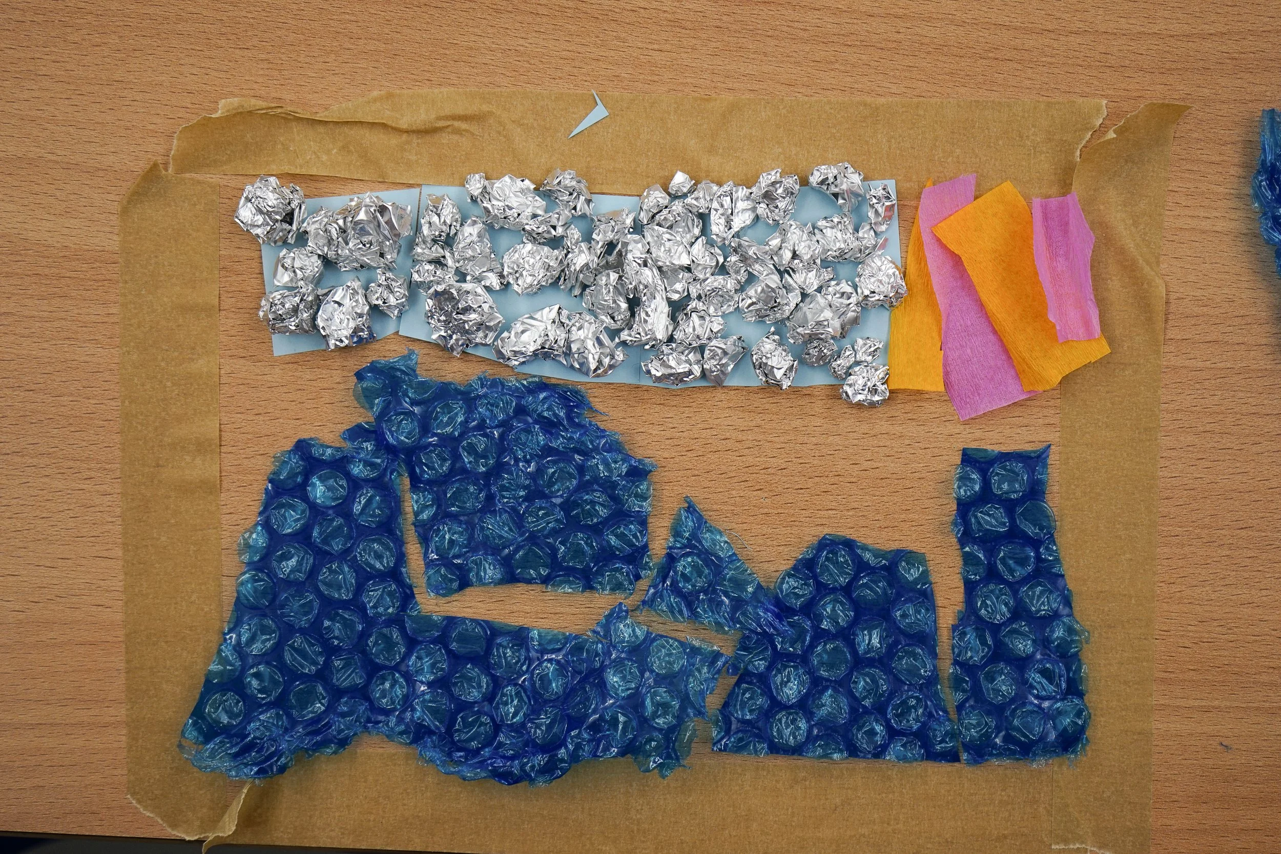 Colorful textured art piece made from torn paper and aluminum foil arranged on a wooden surface.