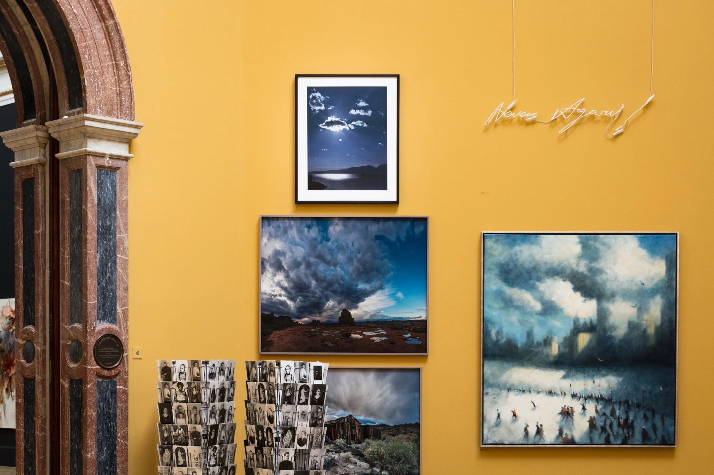 Gallery wall with framed landscape photos of skies and mountains, yellow wall, and a neon sign that reads 'Awards Gallery.' Photographs of black-and-white portraits are displayed underneath.