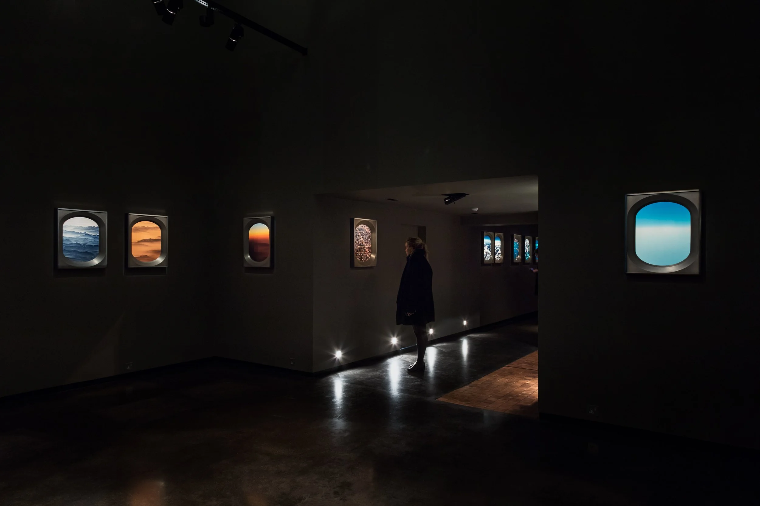 A person is standing in an art gallery with framed landscape photographs hanging on the walls. The room is dimly lit with small spotlights highlighting the artwork.