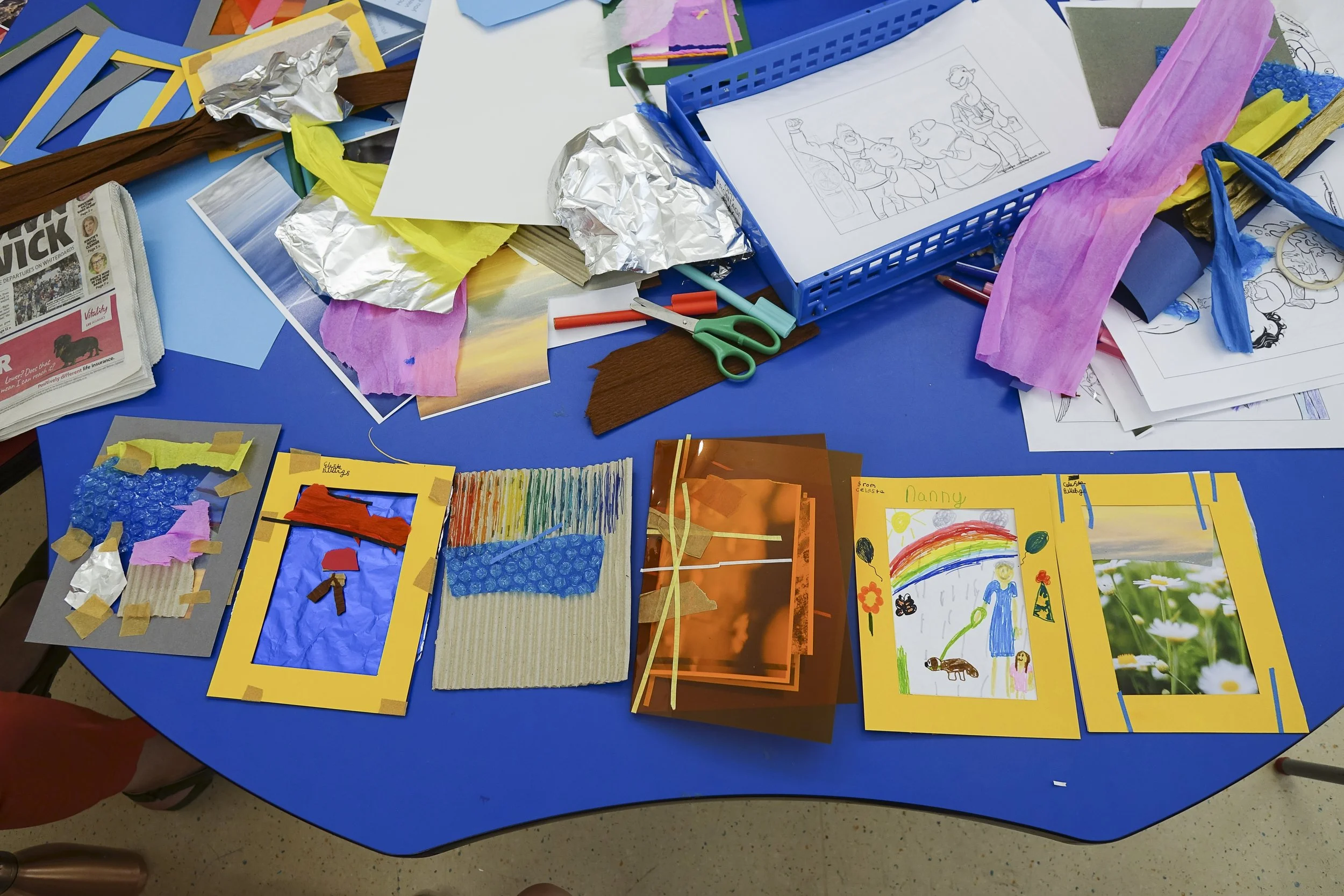 Colorful children's artwork, crafts, and drawings scattered on a blue table, including pictures of rainbows, animals, and people, with craft materials like tissue paper, scissors, and paper scraps.