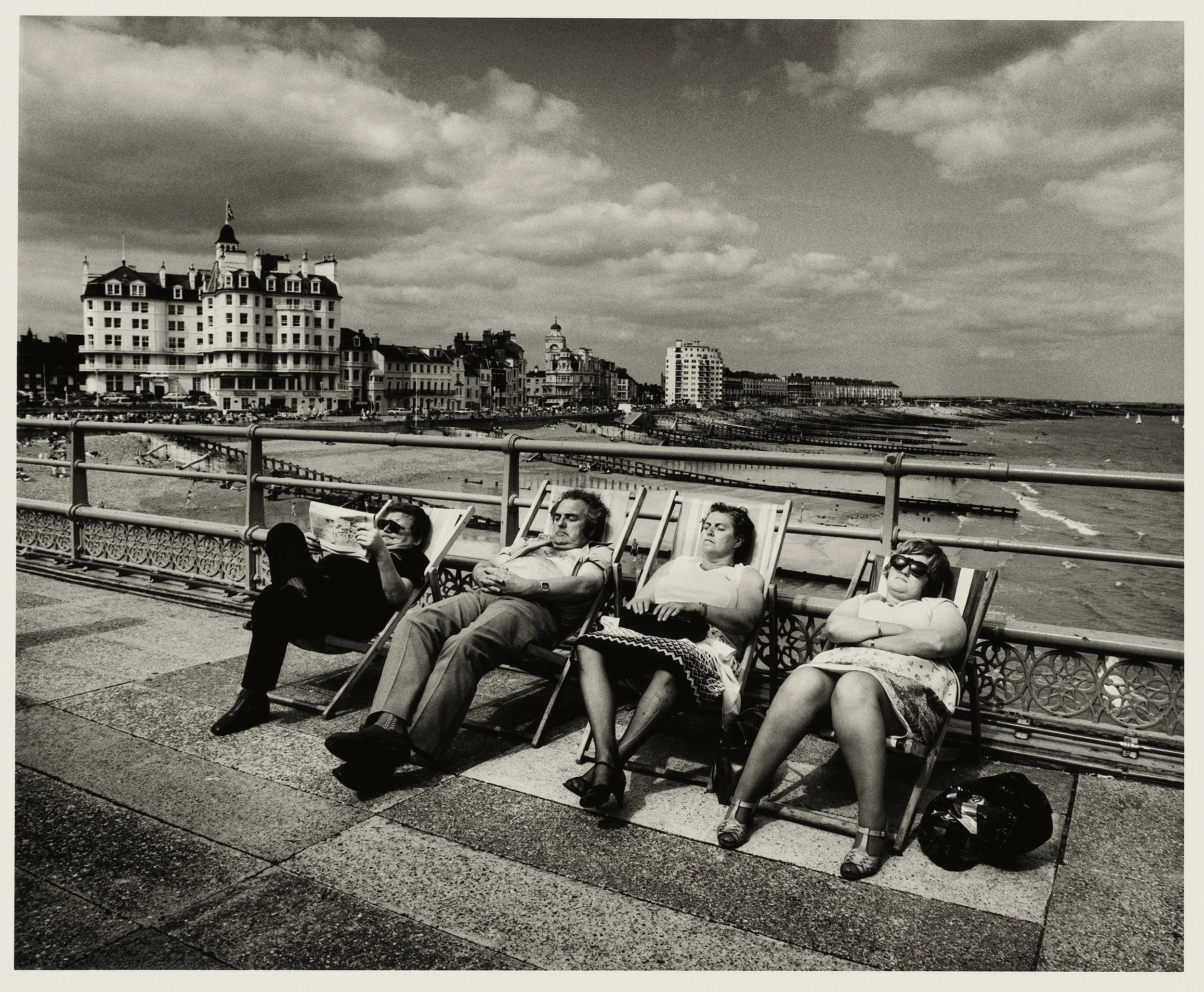 People relaxing on lounge chairs on a seaside promenade with buildings and cloudy sky in the background, black and white photograph.