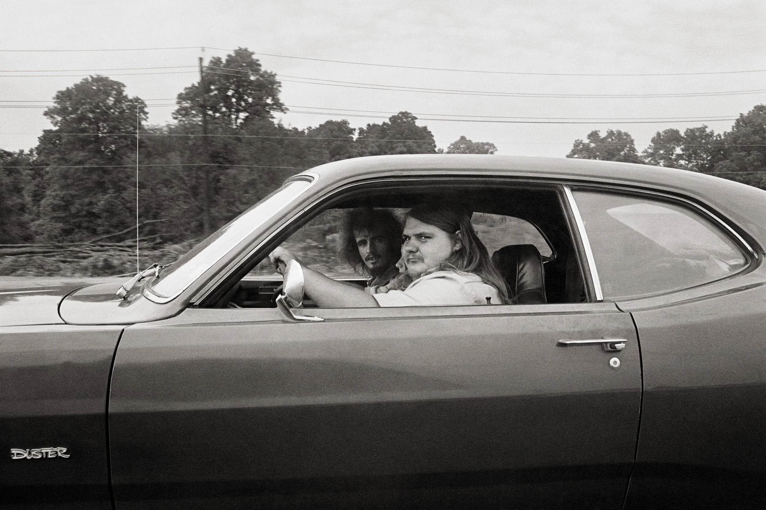 A black and white photo of two people inside a vintage Datsun Duster car on a highway with trees and power lines in the background.