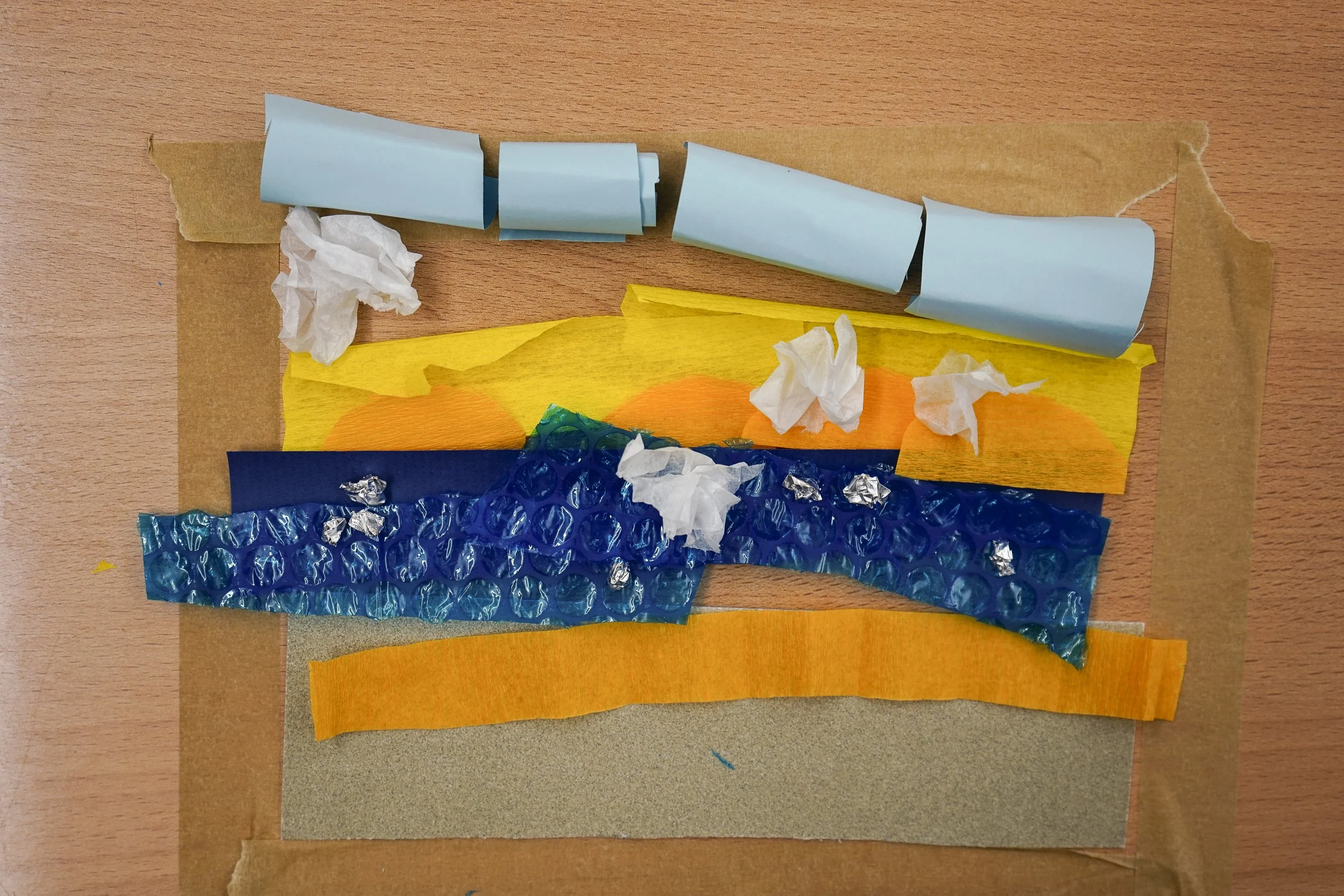 A collage of multiple pieces of colored paper and tissue paper on a wooden surface, including blue, yellow, orange, and dark blue textured materials.