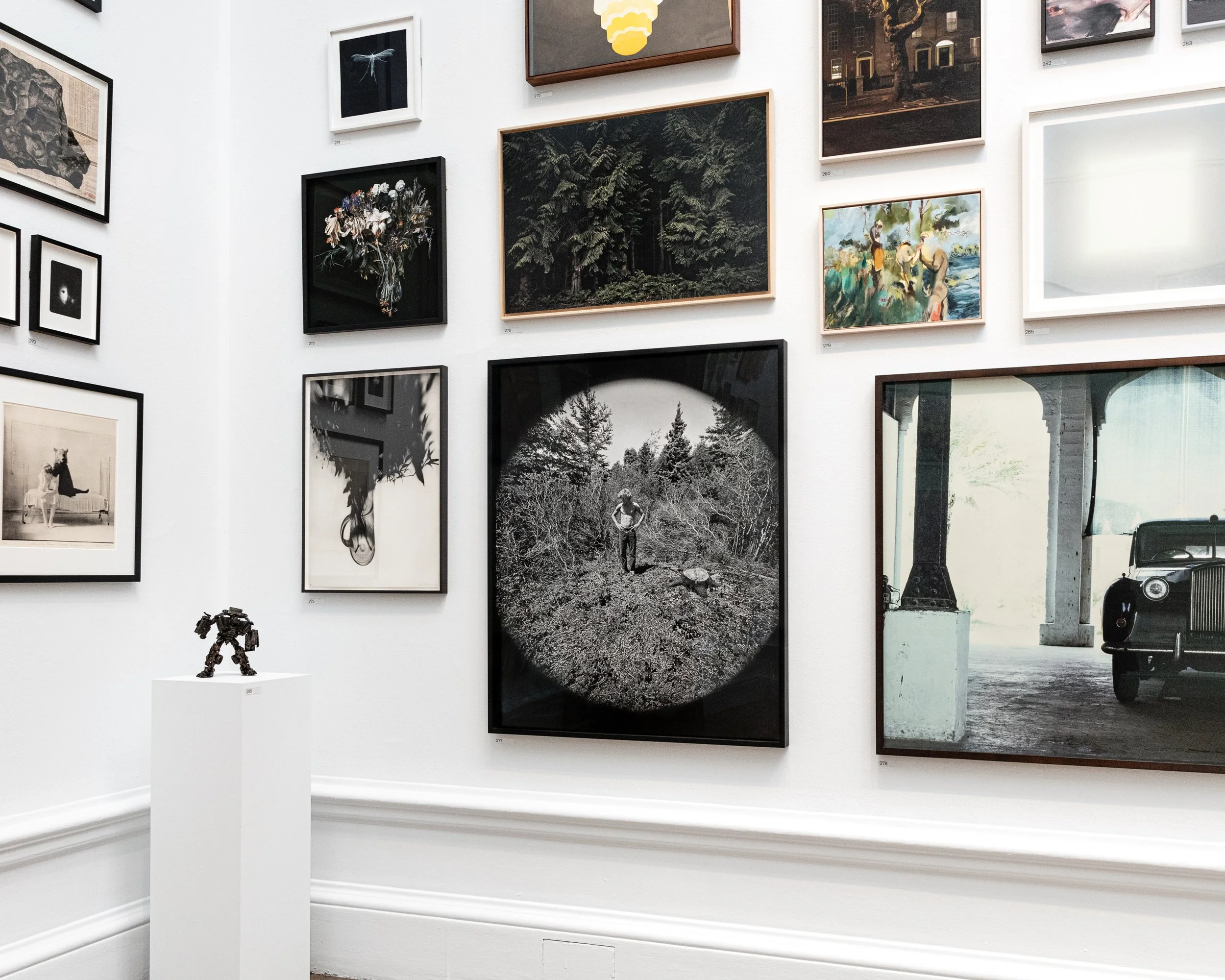 An art gallery wall displaying various framed artworks including photographs, illustrations, and paintings, with a small sculpture of a robot on a white pedestal in front of the wall.