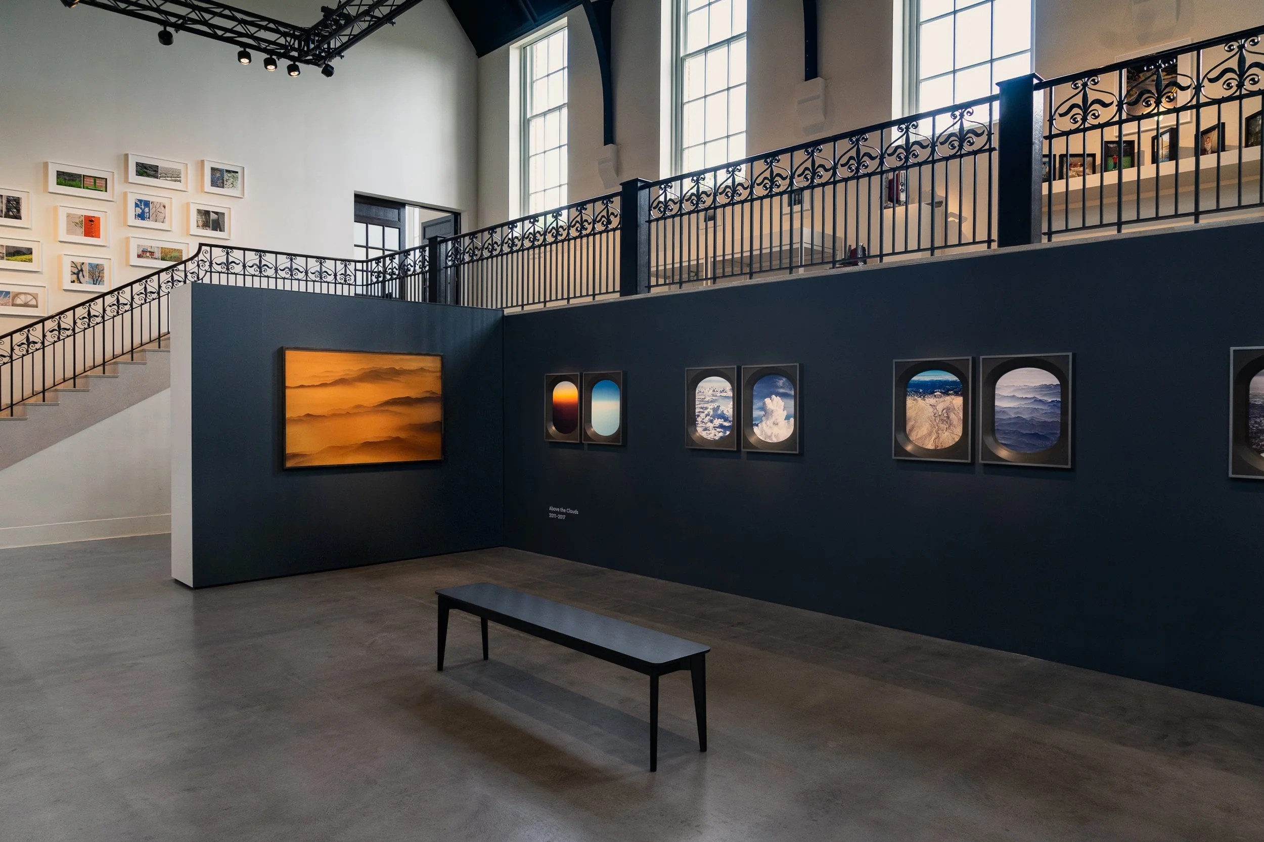 An art gallery displaying photographs and landscape paintings on walls, with a staircase, large windows, and a small black bench in the foreground.