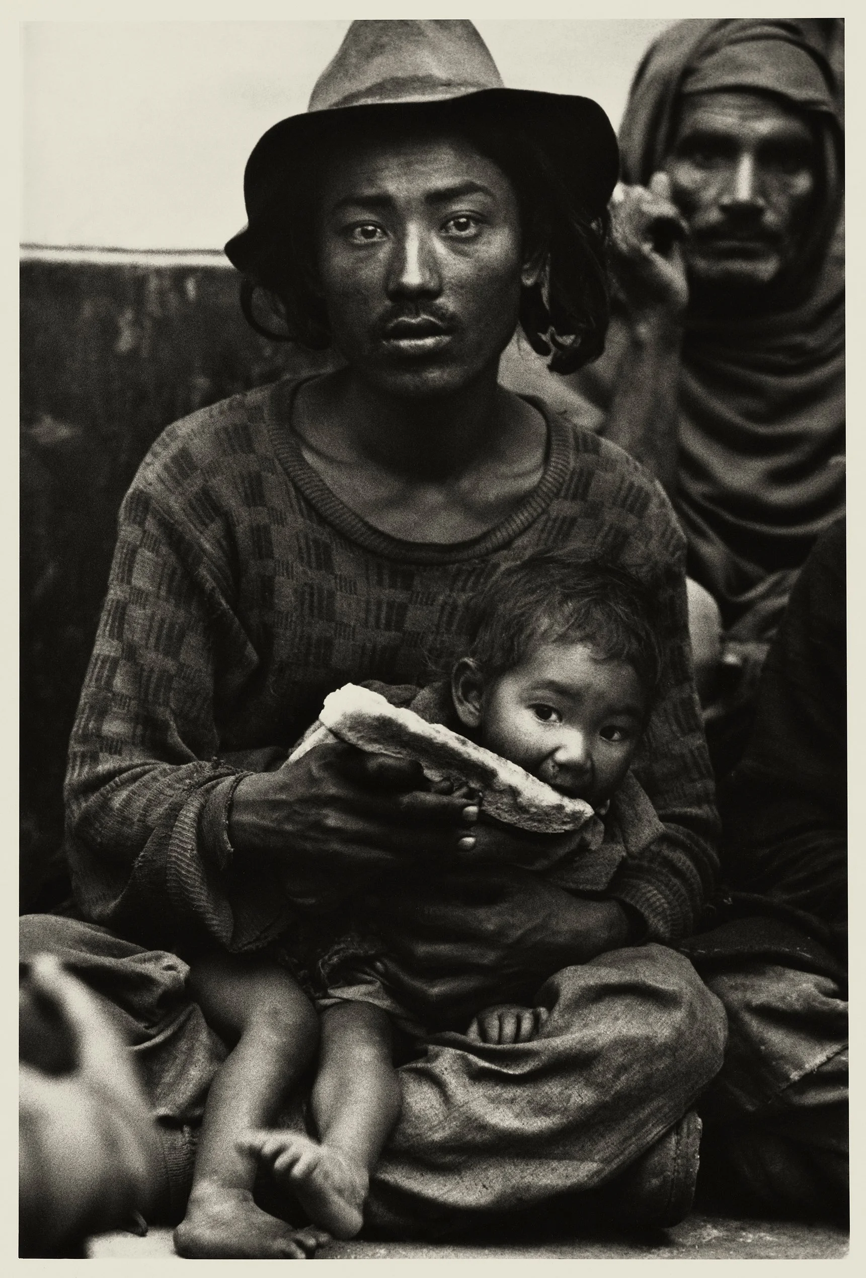 Black and white photo of a woman with a serious expression, wearing a hat, holding a child wrapped in cloth, sitting on the ground, with others in the background.