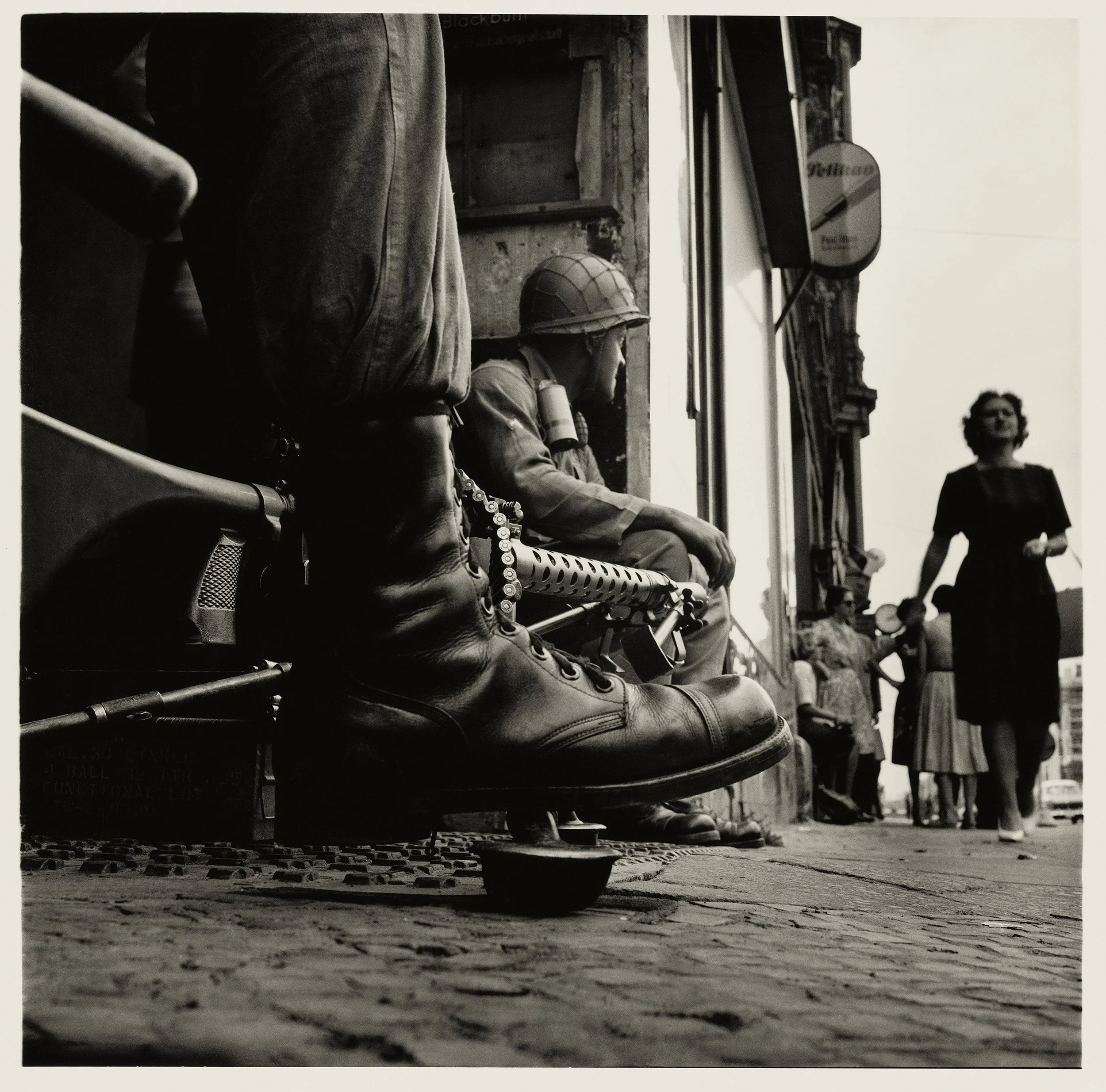 Black and white photograph showing a street scene with a row of women in vintage clothing, with some wearing hats, standing along the sidewalk, and a dog lying on the cobblestone pavement in the foreground.