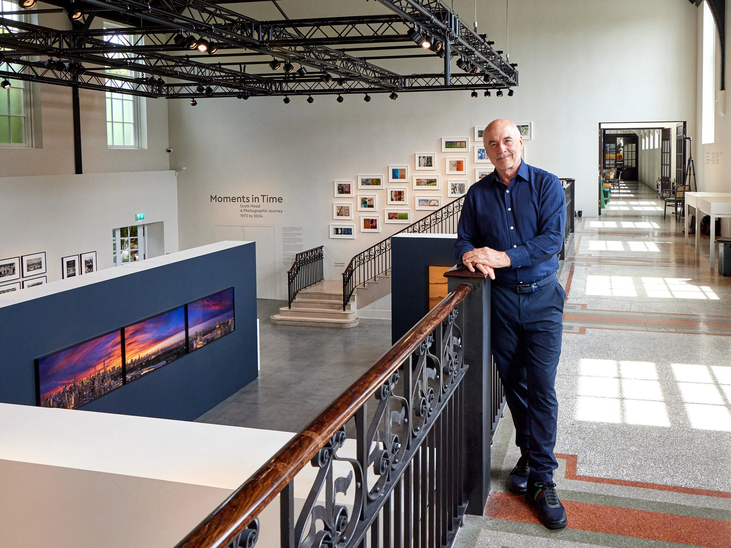 A middle-aged man leaning on a wooden railing inside an art gallery with white walls and multiple framed photographs displayed on the wall. The gallery features a staircase, large windows, and a high ceiling with a metal truss structure and spotlights, showcasing an exhibit titled 'Moments in Time' by Scott Mead.