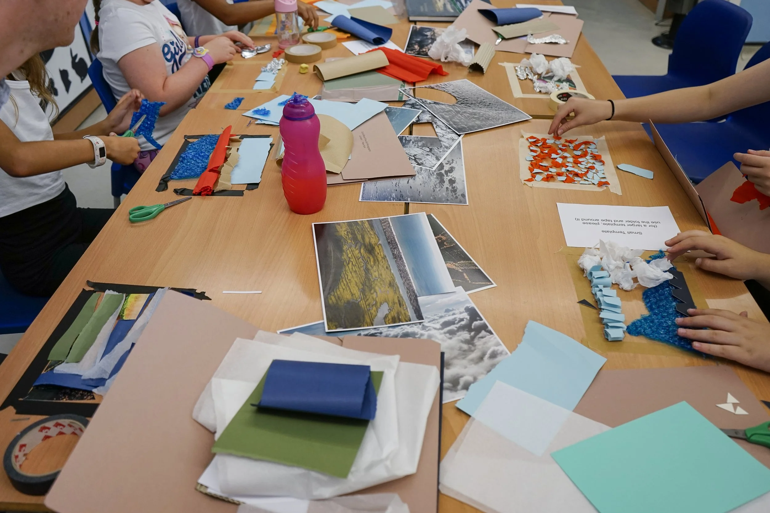 A group of children at a crafting table working on art projects with colored foam, paper, and photographs scattered on the table, with some children holding scissors and glue.