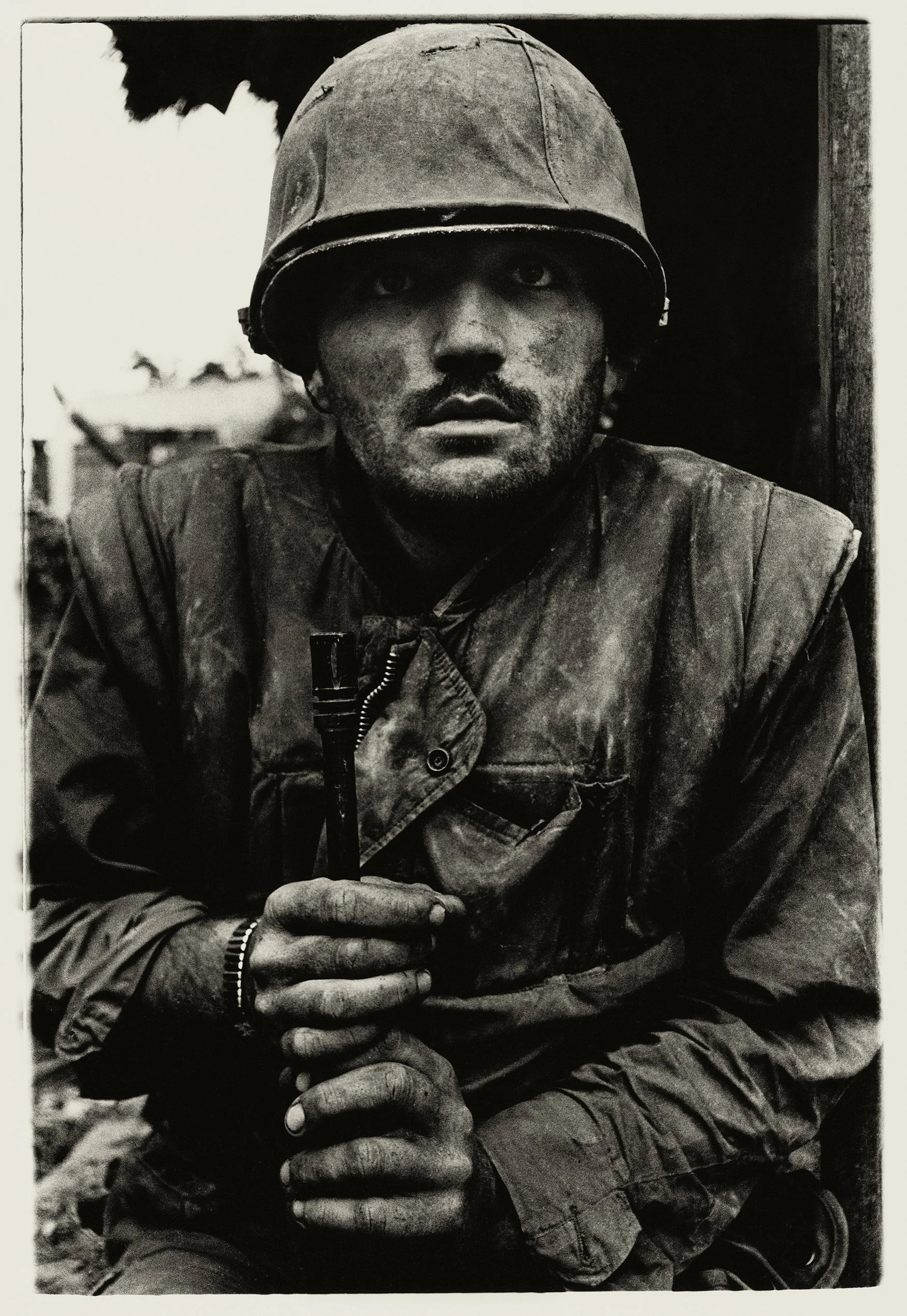 A black-and-white photo of a soldier wearing a helmet, with his hands clasped together, holding a rifle, looking directly at the camera.