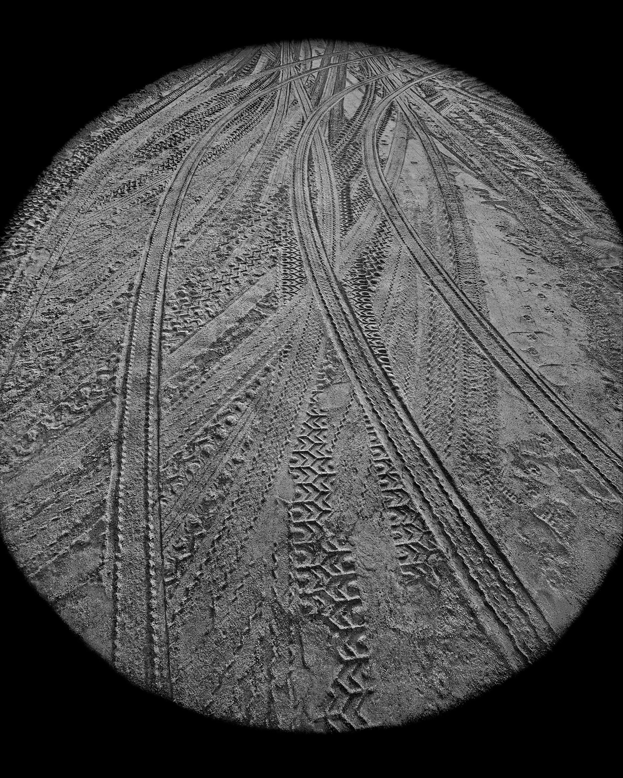 Close-up view of tire tracks and footprints in the sand at night, illuminated by a flashlight or spotlight.