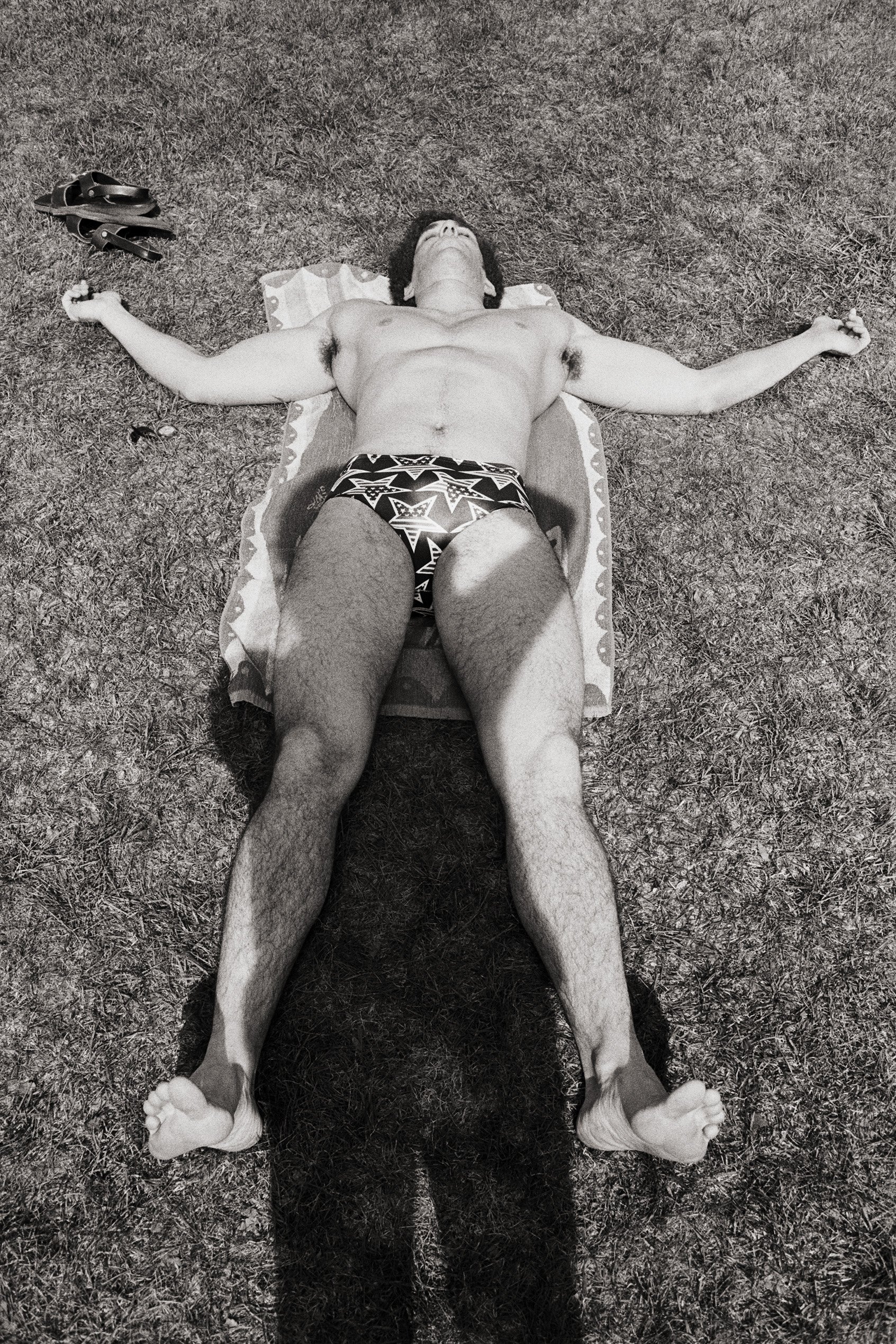 A shirtless man with curly hair wearing patterned swim trunks lies on a towel on the grass with arms and legs spread out.