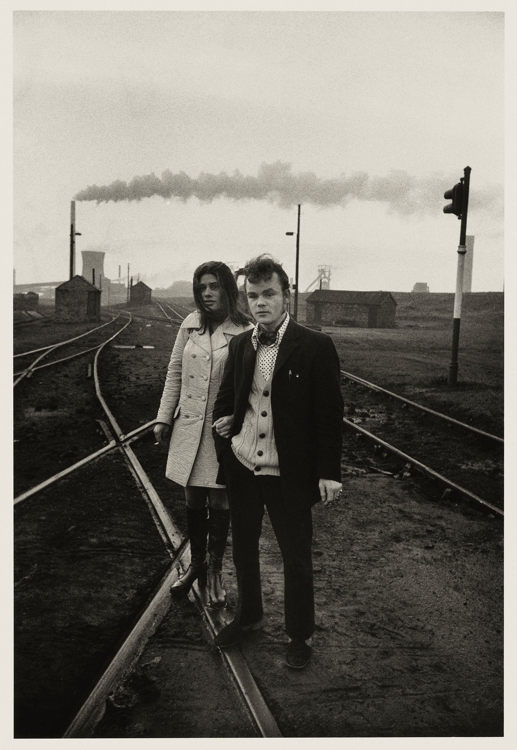 Two young people standing on railroad tracks outdoors with industrial buildings and smoke in the background, black and white photograph