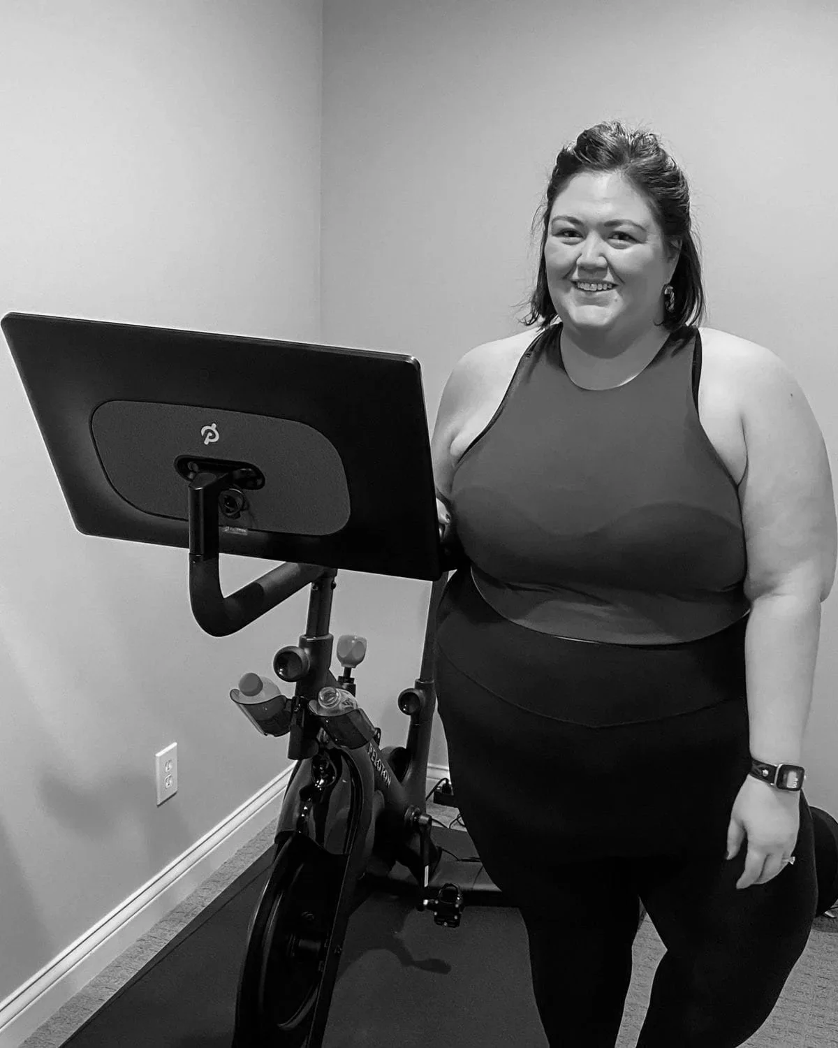 Smiling woman standing beside a Peloton exercise bike in a room