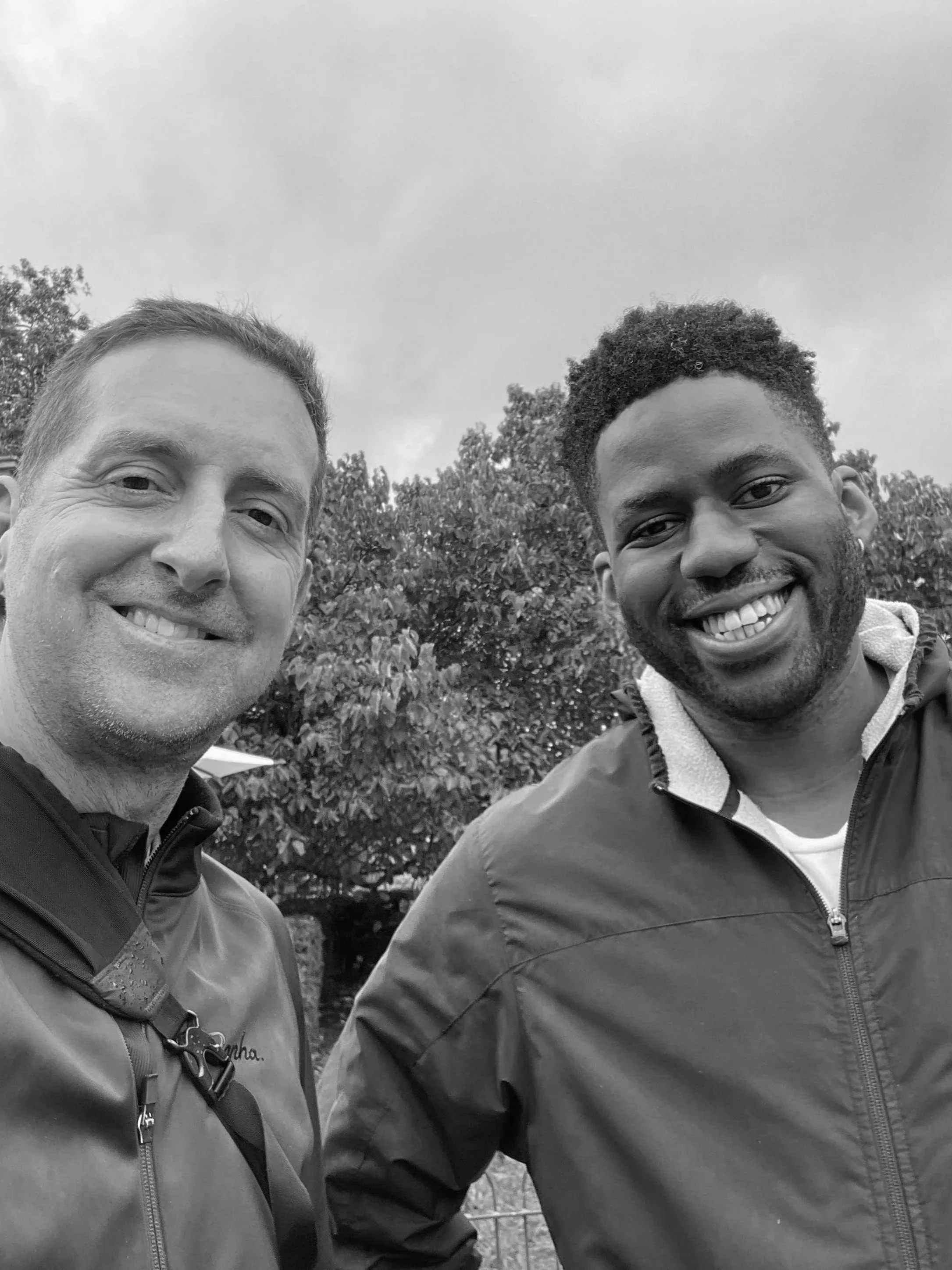 Black-and-white outdoor selfie of two smiling men in jackets with trees in the background.