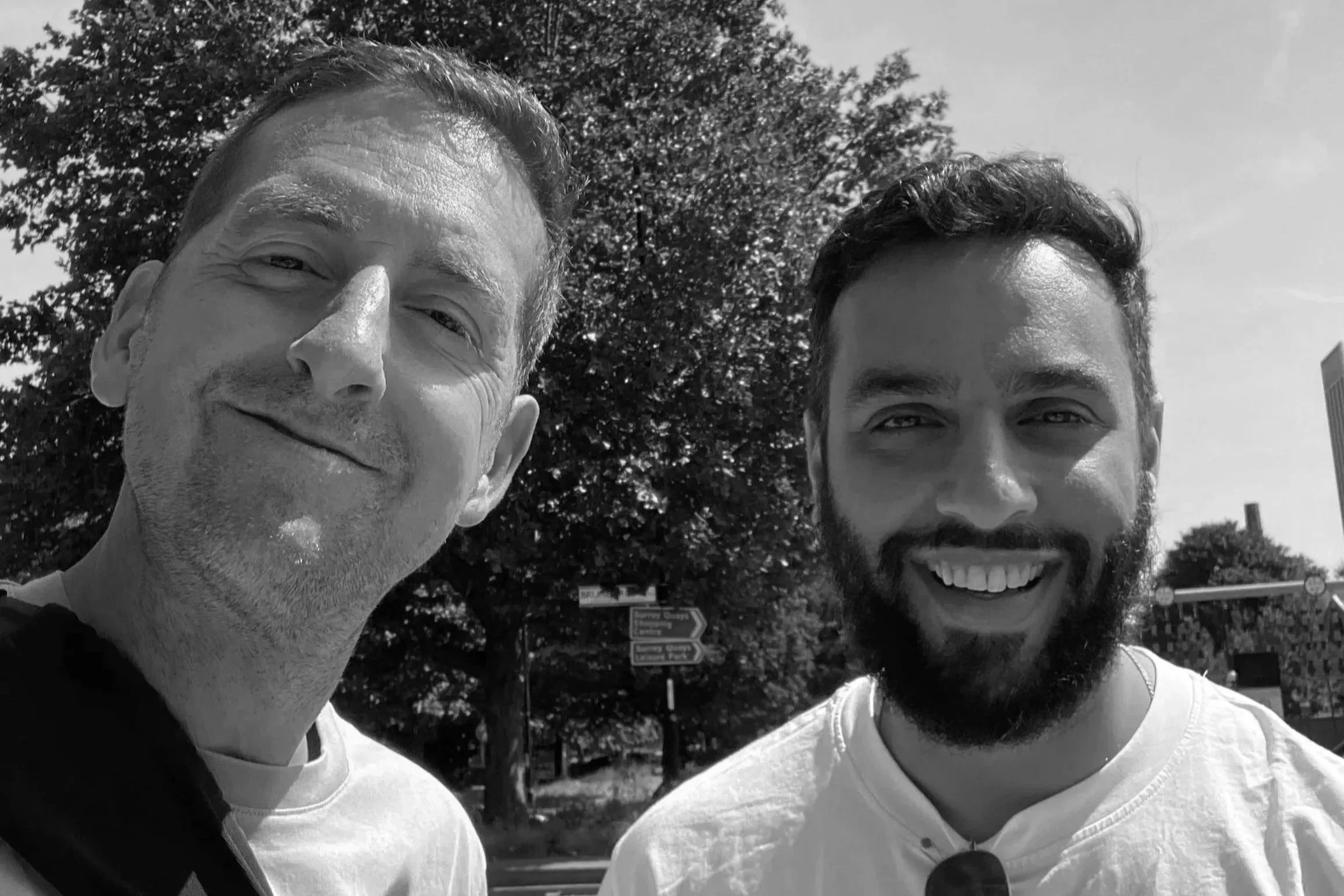 Black-and-white selfie of two smiling men outdoors with trees and directional signs in the background