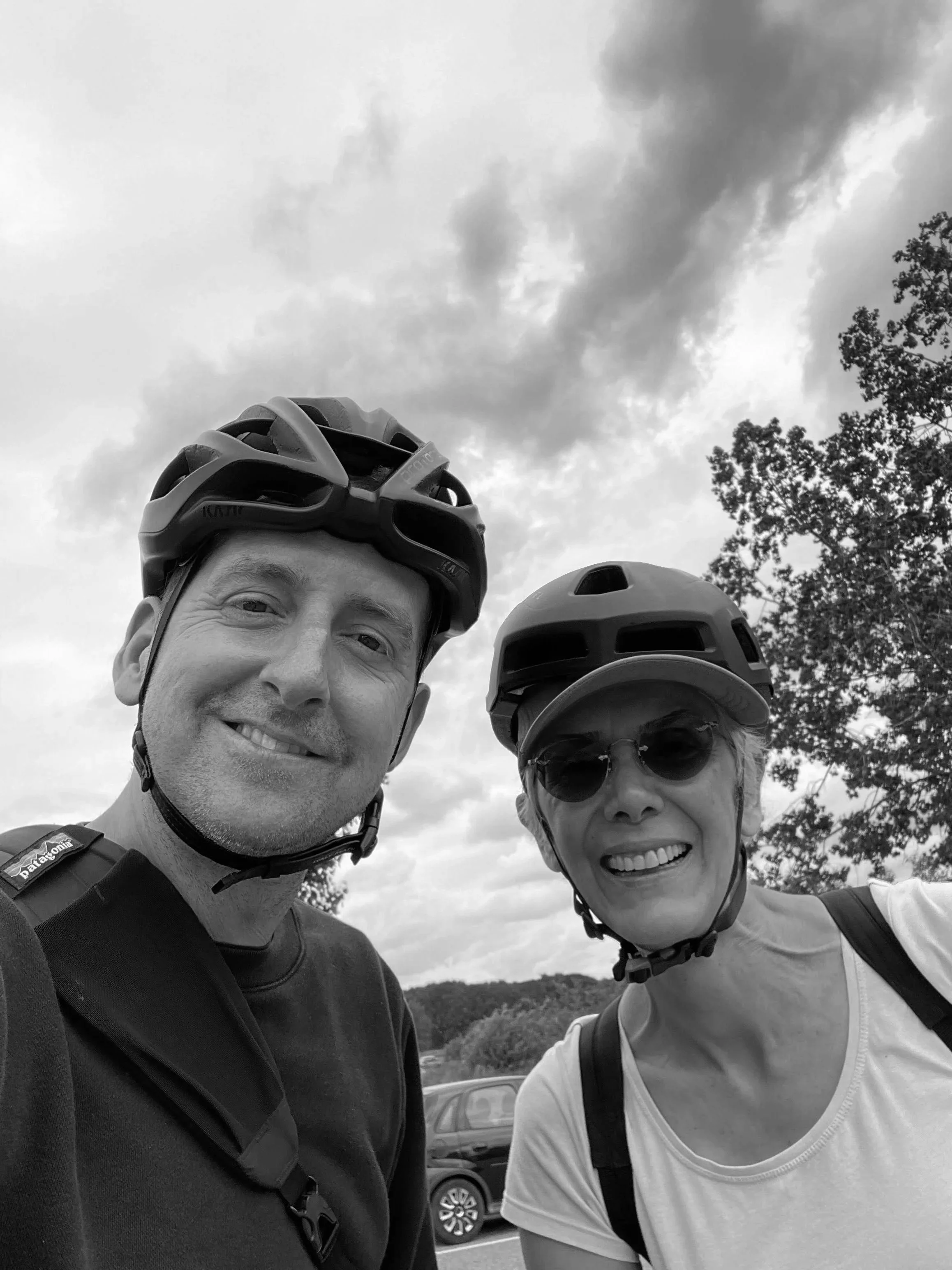 Black-and-white selfie of two smiling cyclists wearing helmets and sunglasses outdoors beneath a cloudy sky