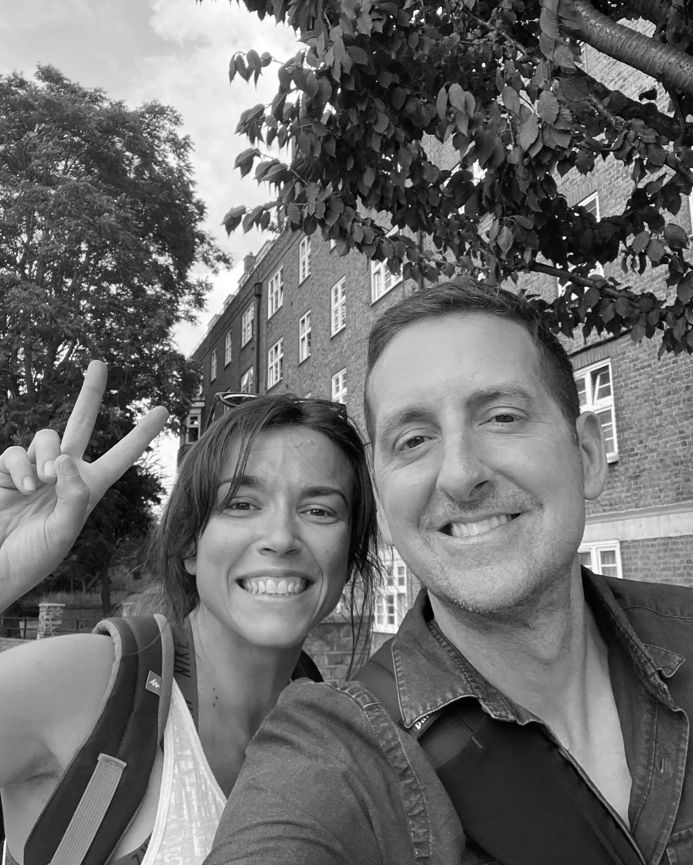 Black-and-white selfie of two smiling people outdoors in front of a brick building and leafy trees, with a hand making a peace sign.