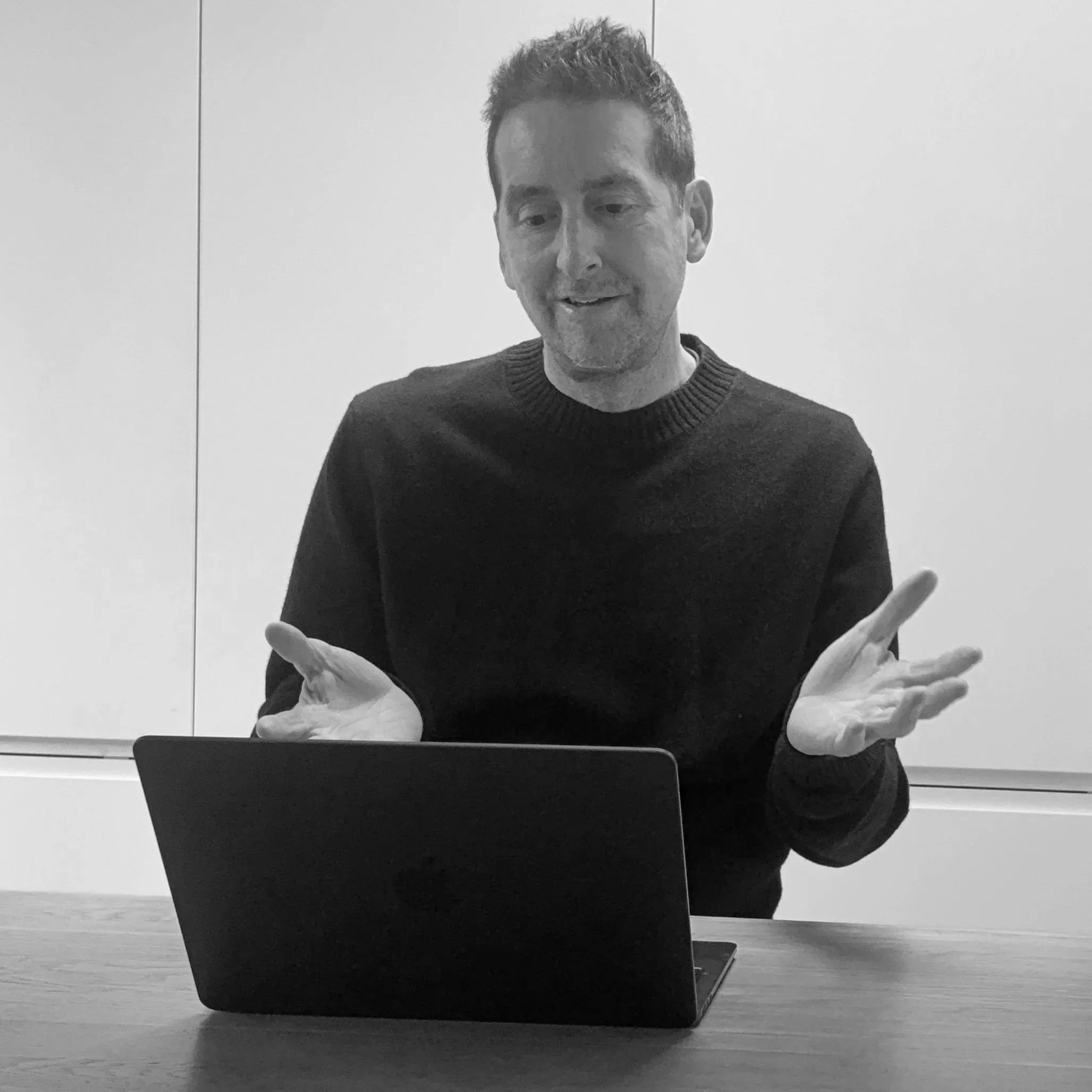 Man in a dark sweater gesturing behind an open laptop at a table