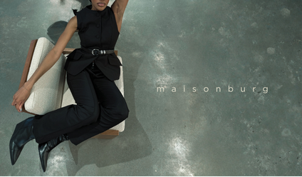 Person lying on their back on a carpeted floor next to a chair, wearing a black outfit, holding a large white bag, with "maisonburg" written on the floor.
