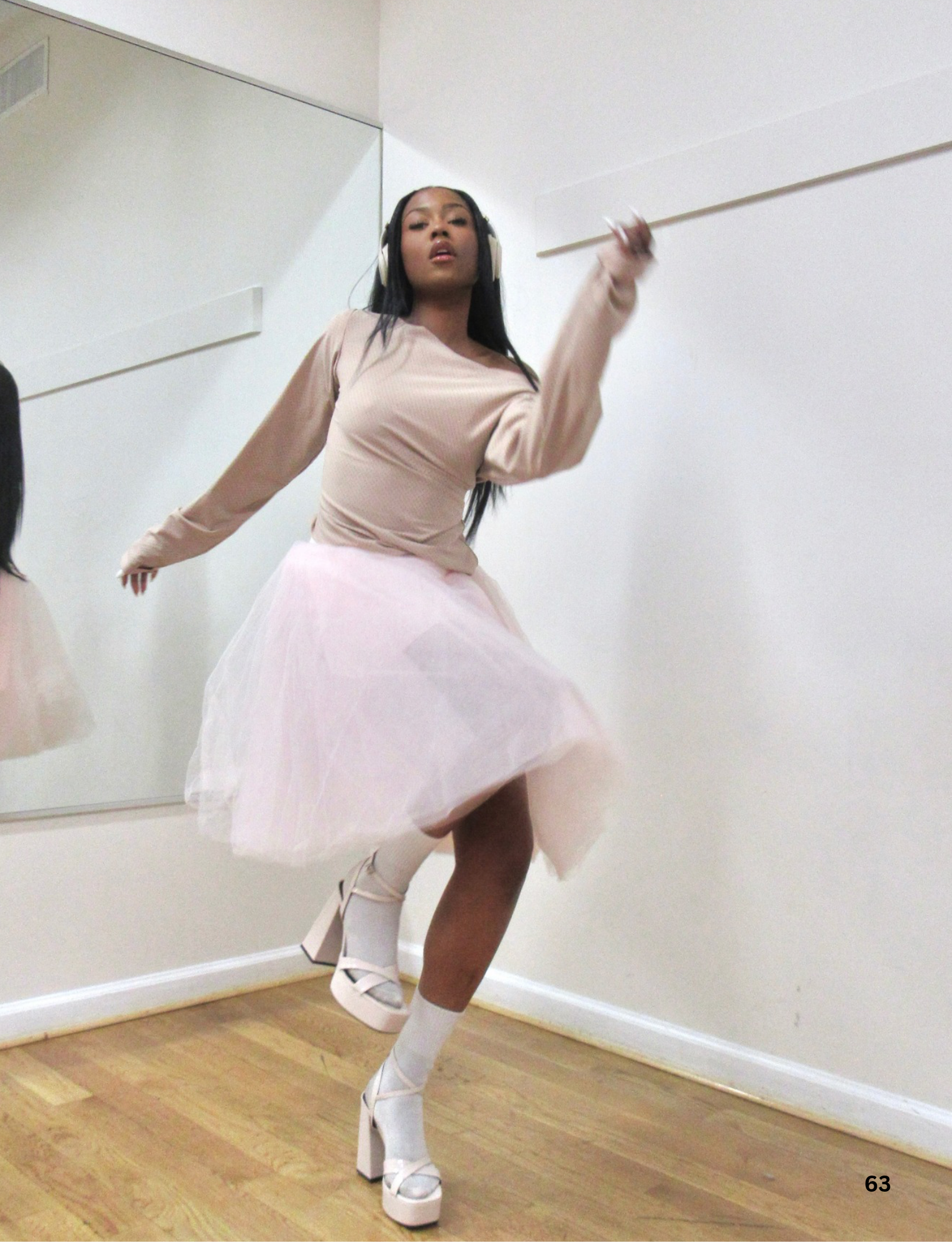 A woman dancing in a room with white walls and a wooden floor, wearing a light pink dress, white platform shoes, and large headphones.