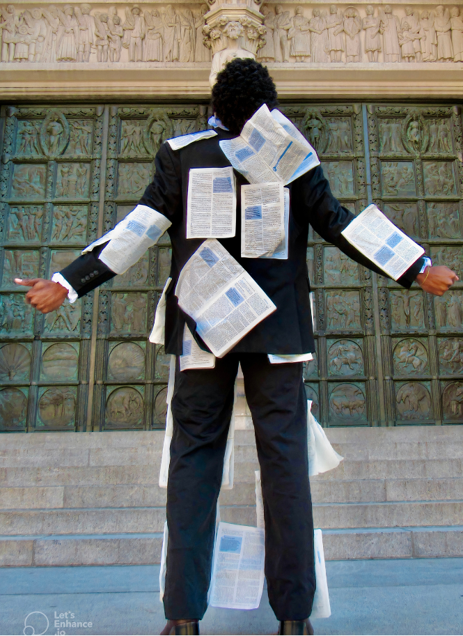 A person dressed in a black suit with newspapers pinned all over the back, front, and sleeves, standing with arms outstretched in front of a large decorative metal structure in an outdoor setting.