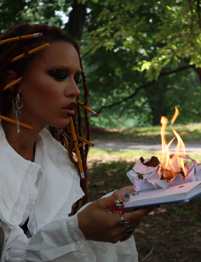 A woman with red dreadlocks and dark makeup is holding a book with crumpled pages that are on fire, in an outdoor setting with green trees in the background.
