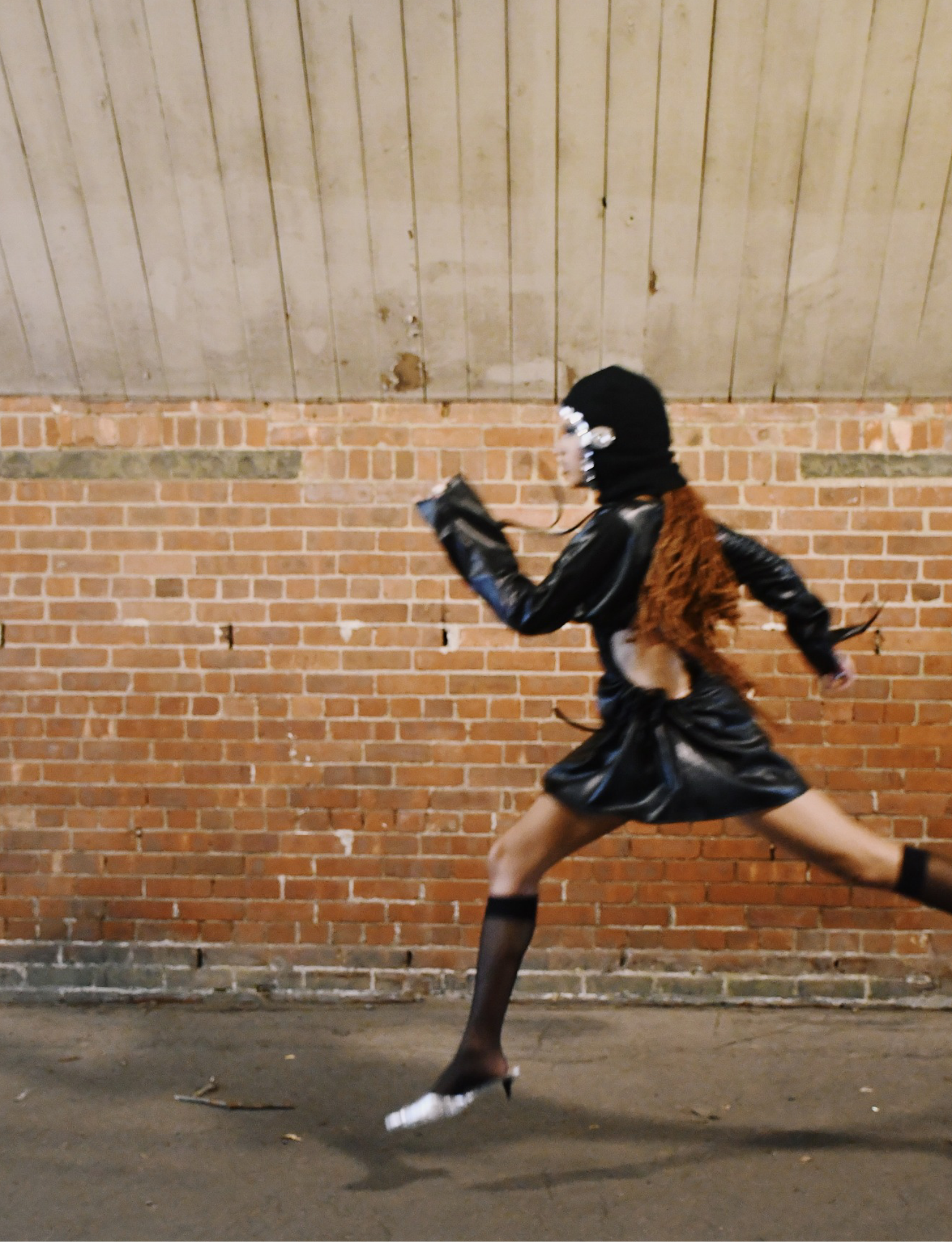 Person dressed in black leather jacket, furry vest, and black skirt running in front of a brick wall and wooden ceiling.