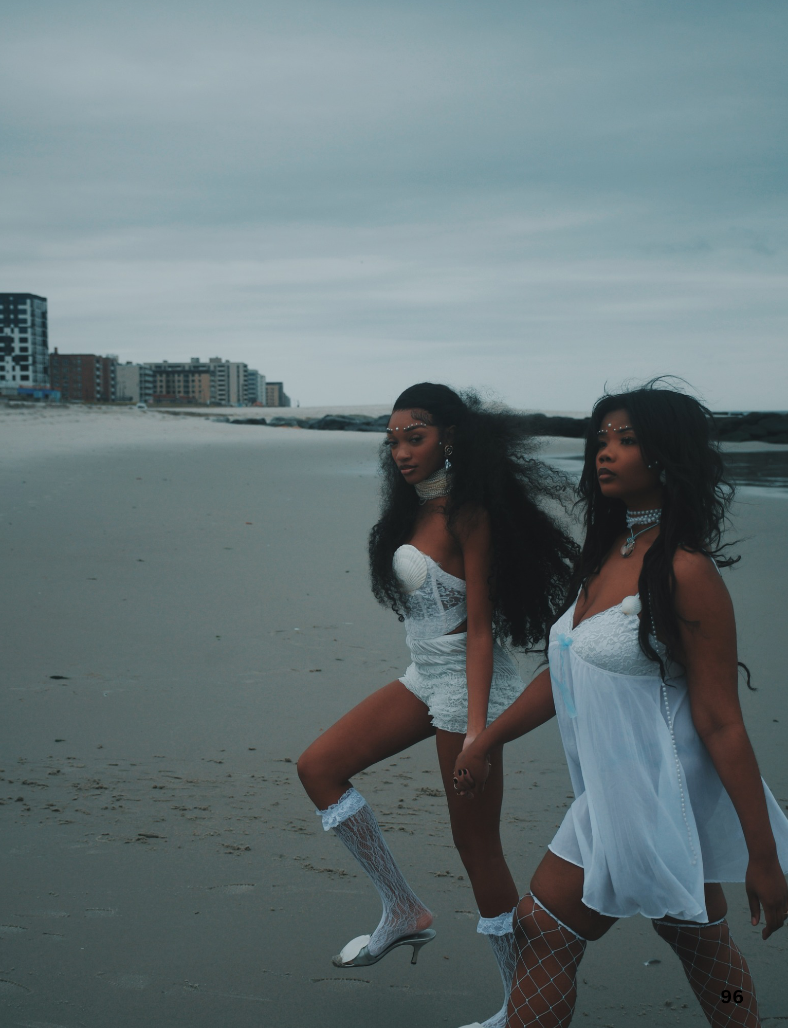 Two women with curly hair and jewelry standing on a beach with city buildings in the background, dressed in white dresses and stockings, posing for a photo.