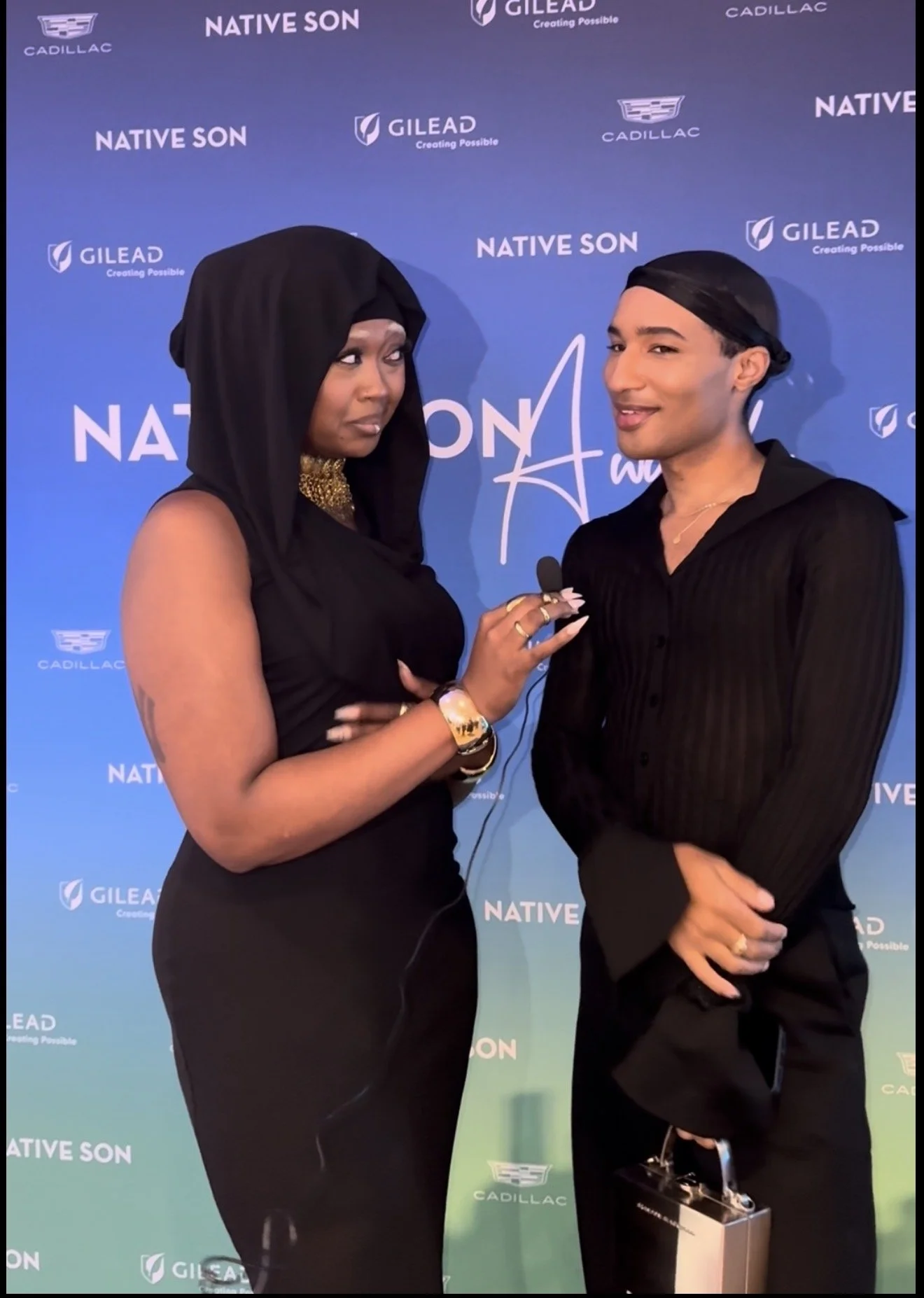 Two people, a woman with dark skin wearing a black dress and black head covering, and a man with light skin wearing a black shirt and head covering, standing in front of a blue backdrop with logos and text. The woman appears to be interviewing the ma
