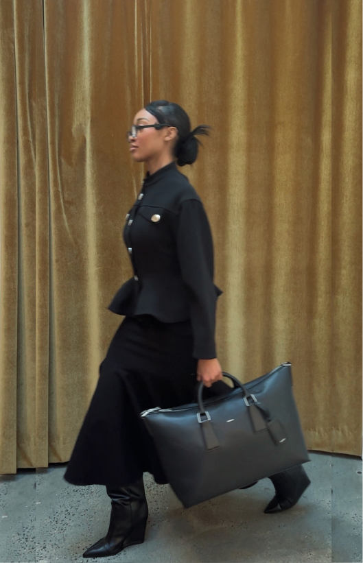 A woman in black business attire, wearing glasses, walking past gold curtain backdrop while holding a large black handbag.