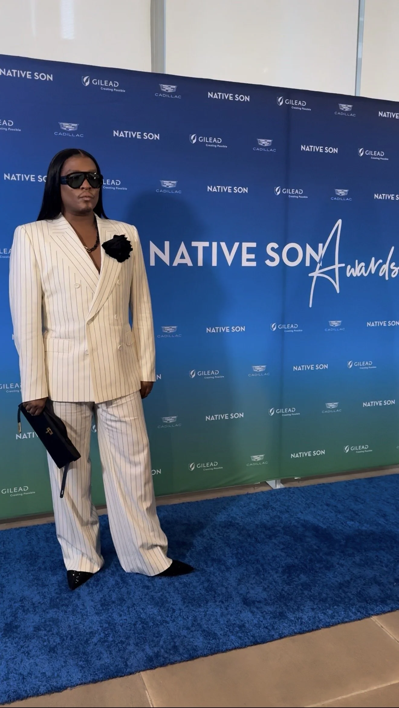 Person standing in front of a blue backdrop with logos and text for Native Son Awards and Gilead, dressed in white pinstripe suit, wearing sunglasses and holding a clutch.