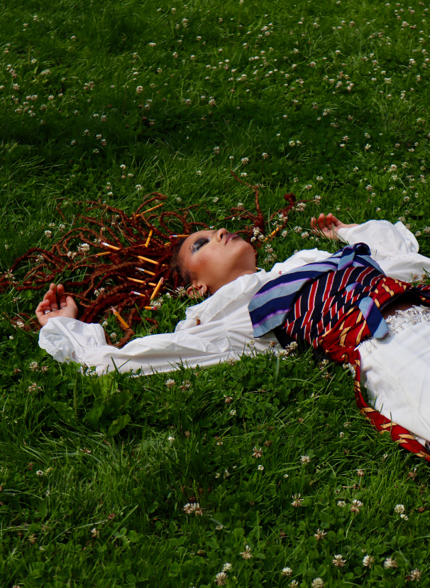 Woman lying on grass with colorful hair and cigarettes in her hair, wearing a white shirt and patterned skirt.