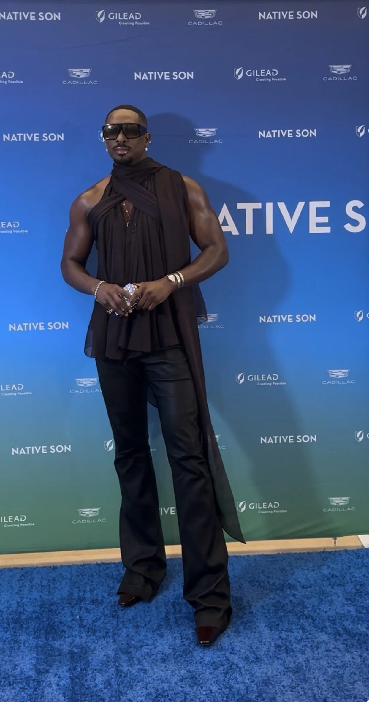 A man standing in front of a blue backdrop with logos of Native Son, Gilead, and Cadillac. He is wearing sunglasses, a sleeveless dark top with fabric draped over his shoulders and a high-slit black pair of pants with pointed shoes.