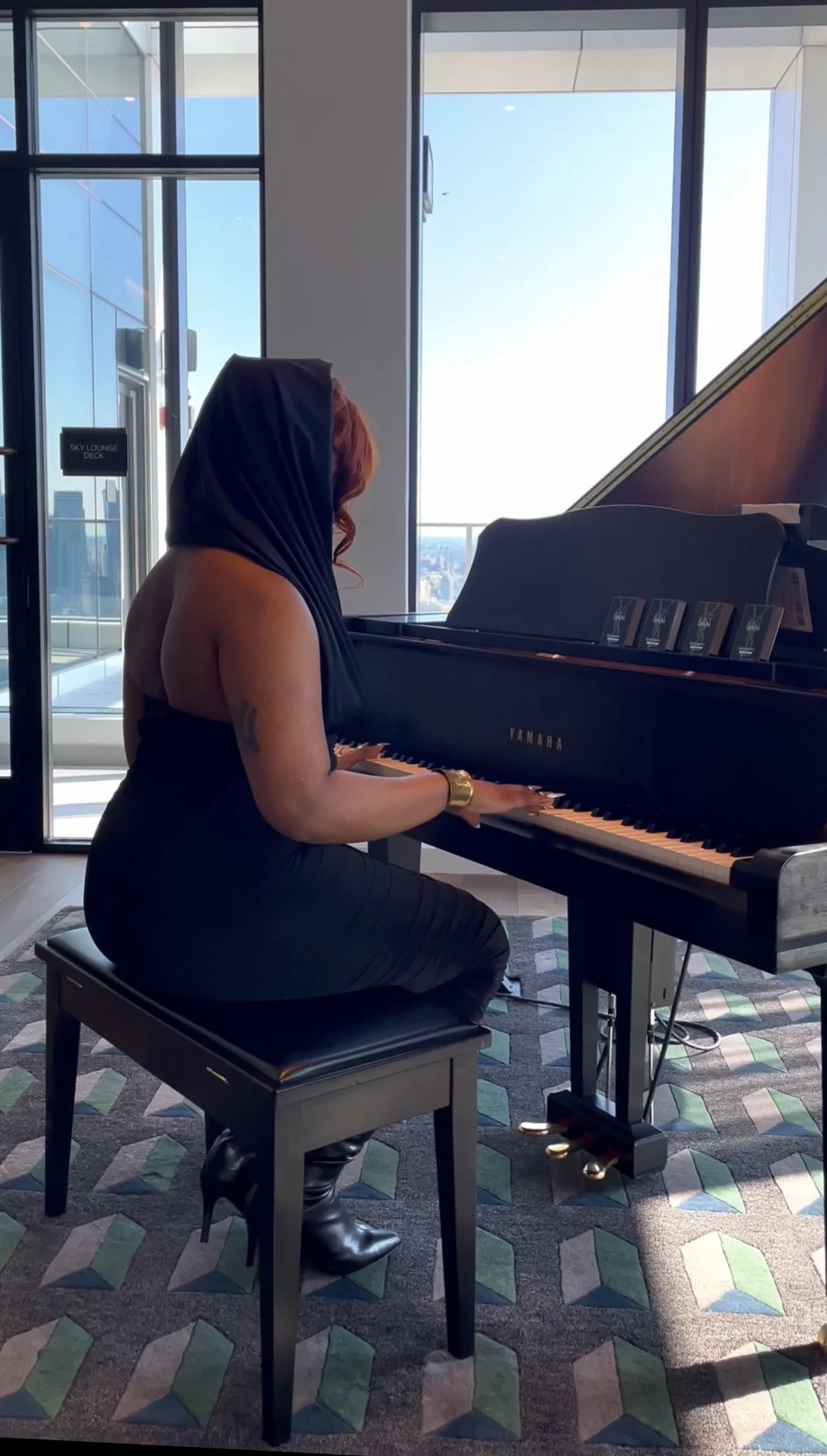 A woman with red hair and a blue headscarf playing a black Yamaha grand piano in a modern room with large windows and a city view.