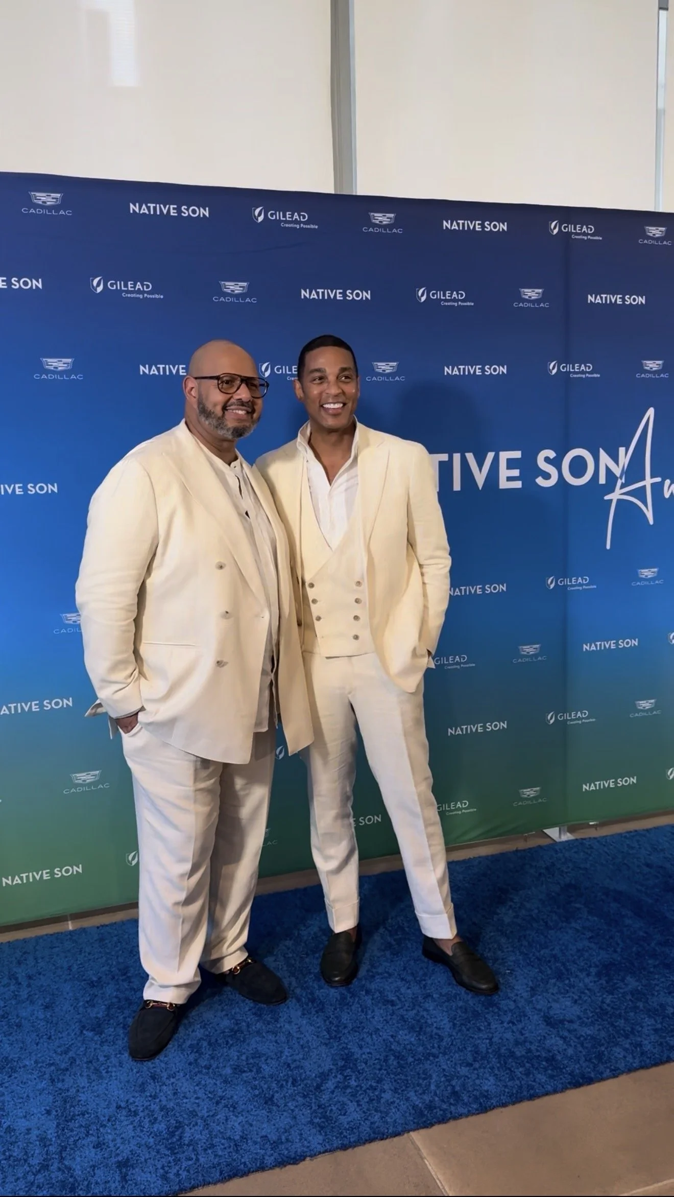 Two men standing in front of a blue backdrop with logos of Cadillac, Gilead, and Native Son, wearing cream-colored suits, smiling at a promotional event.