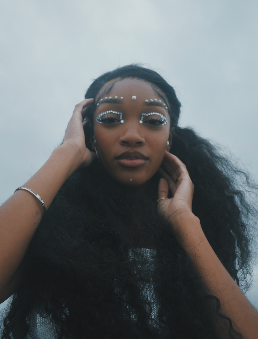 A woman with long, curly black hair and dark skin, wearing intricate white face and eye makeup with rhinestones, looking at the camera with one hand touching her head and the other near her neck, against a cloudy sky background.