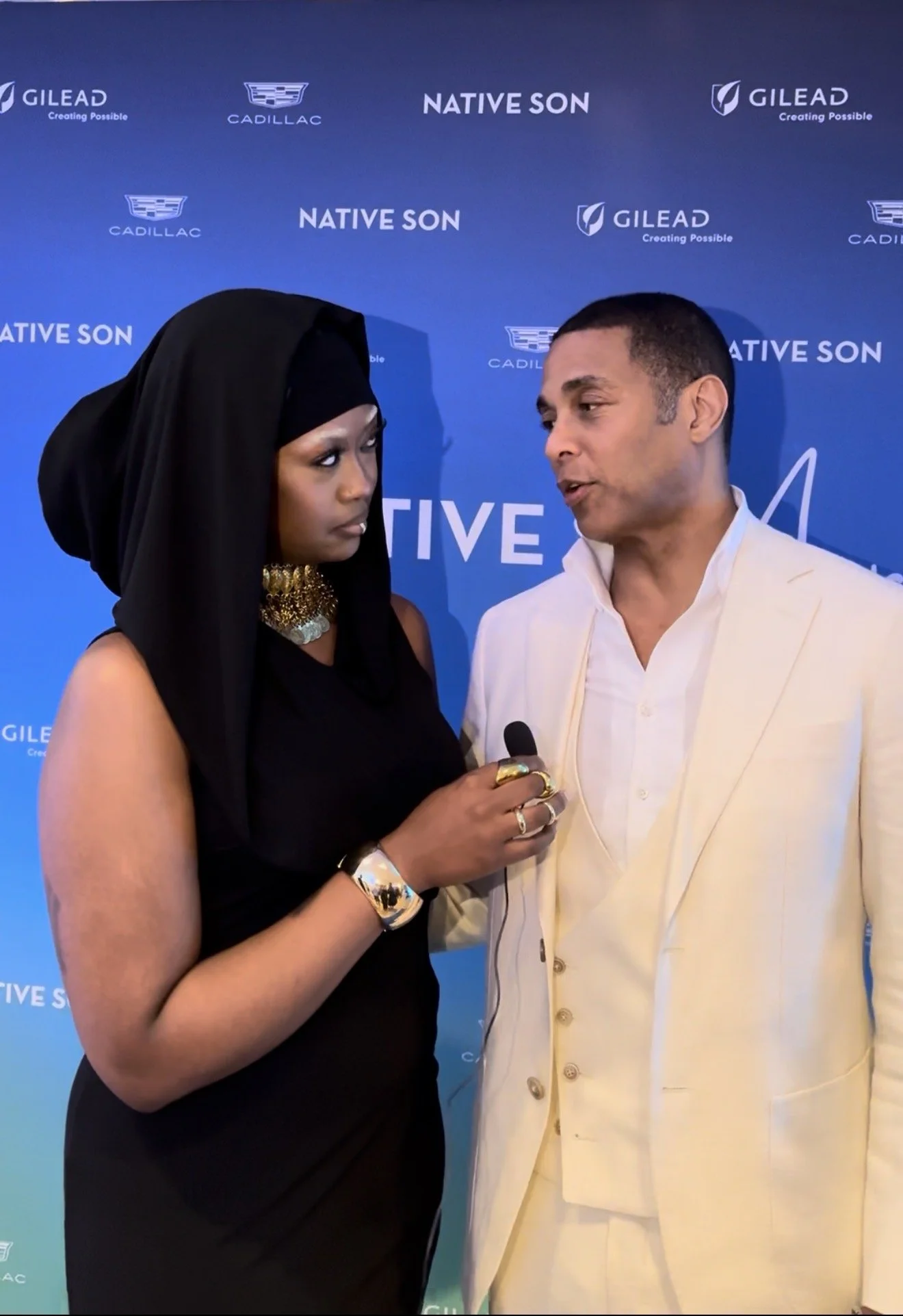Two people, a woman in black and a man in cream-colored suit, are engaged in a conversation against a blue backdrop with logos and the words 'Native Son', 'Gilead', and 'Cadillac'.