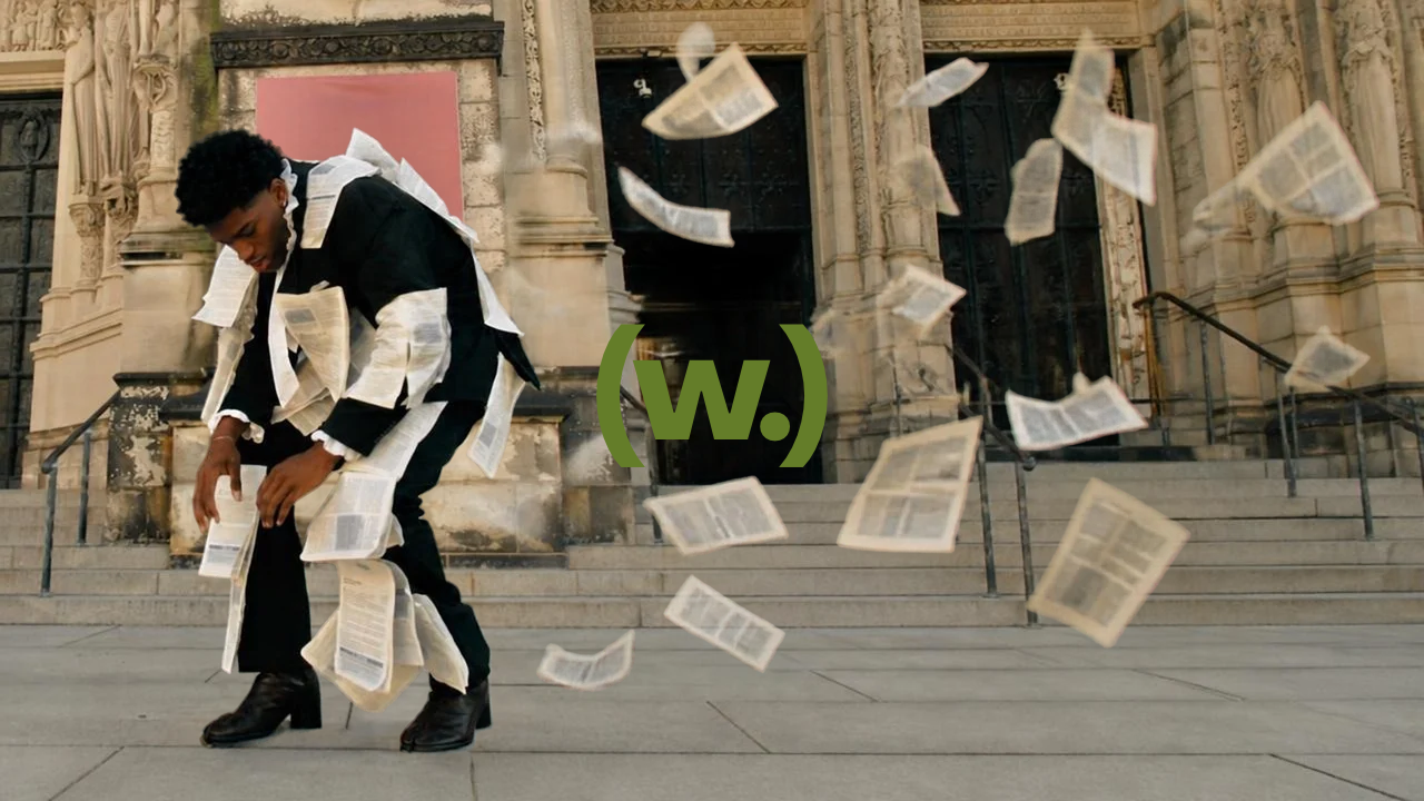 A person dressed in black suit with white paper attached to clothing, bending down on steps outside a historic stone building with columns, as papers fly around in the air.