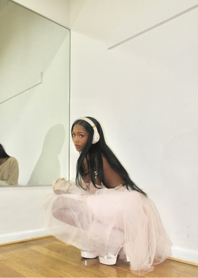 A young woman with long dark hair wearing large white headphones, a sheer black top, and a light pink tulle skirt squatting in a room with a mirror on the wall and wooden floor.